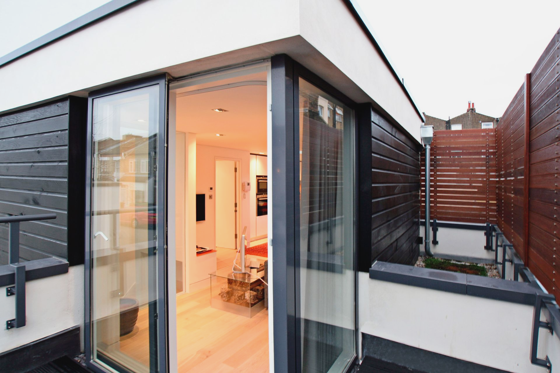 Modern apartment balcony with open glass doors, warm-lit interior, and dark wood privacy screens