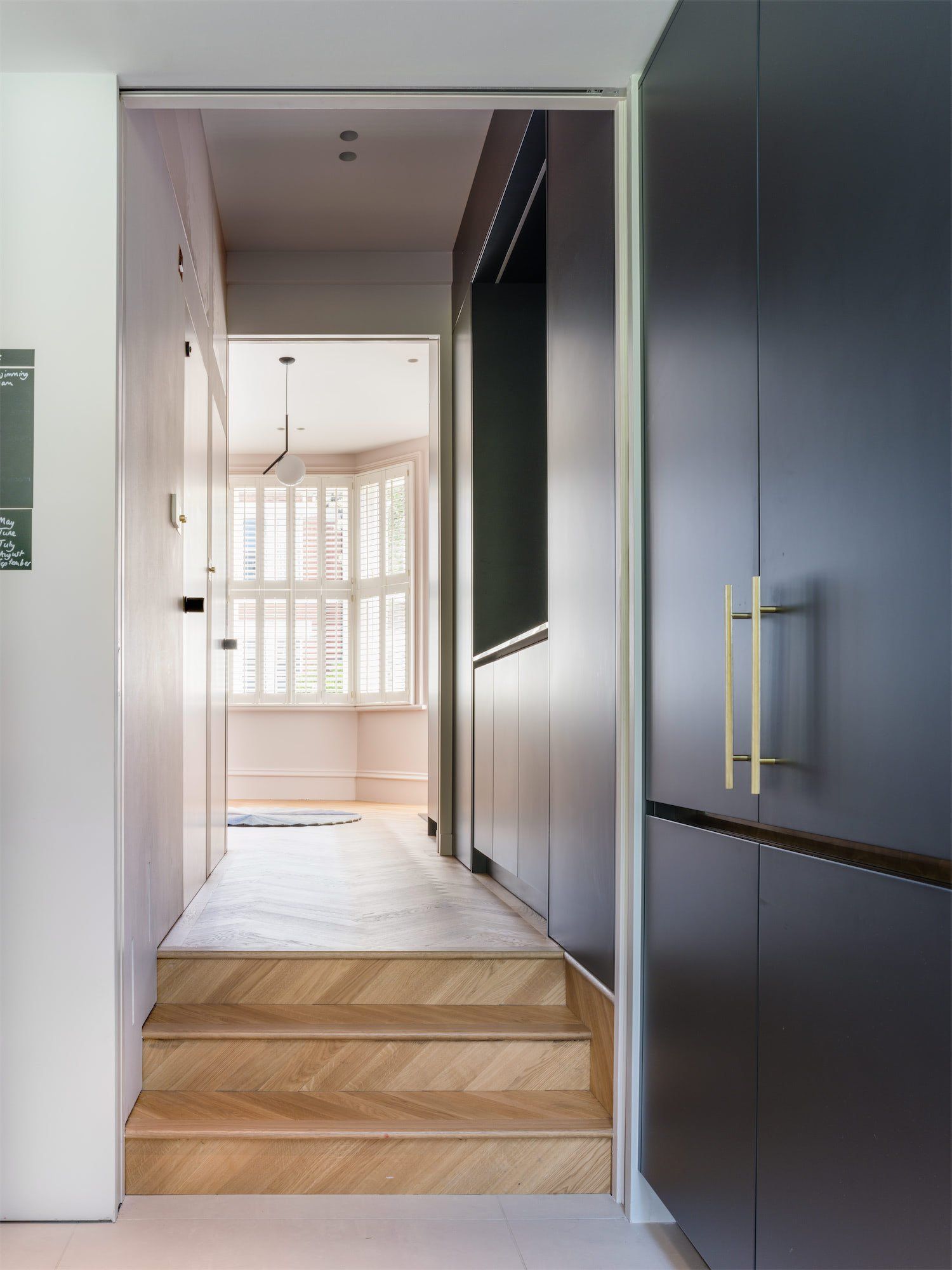 Modern hallway with wooden steps, white walls, and dark built-in cabinets leading to a bright room.