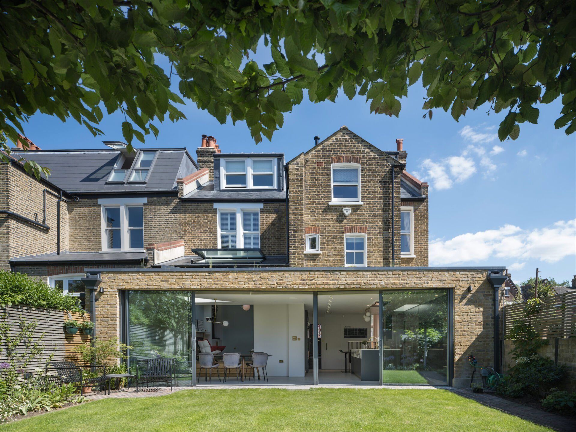 Stone house with solar panels and a covered patio, viewed from a leafy garden under a blue sky