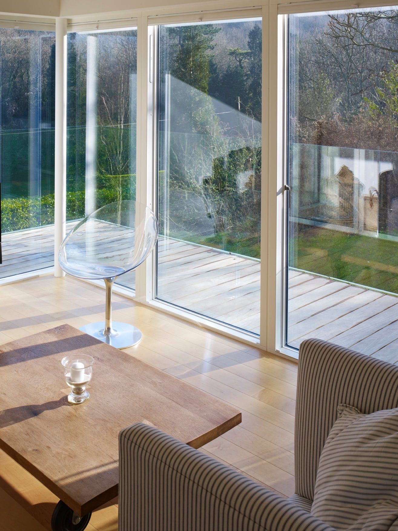Sunlit living room with striped armchair, wooden coffee table, and large glass windows overlooking a garden