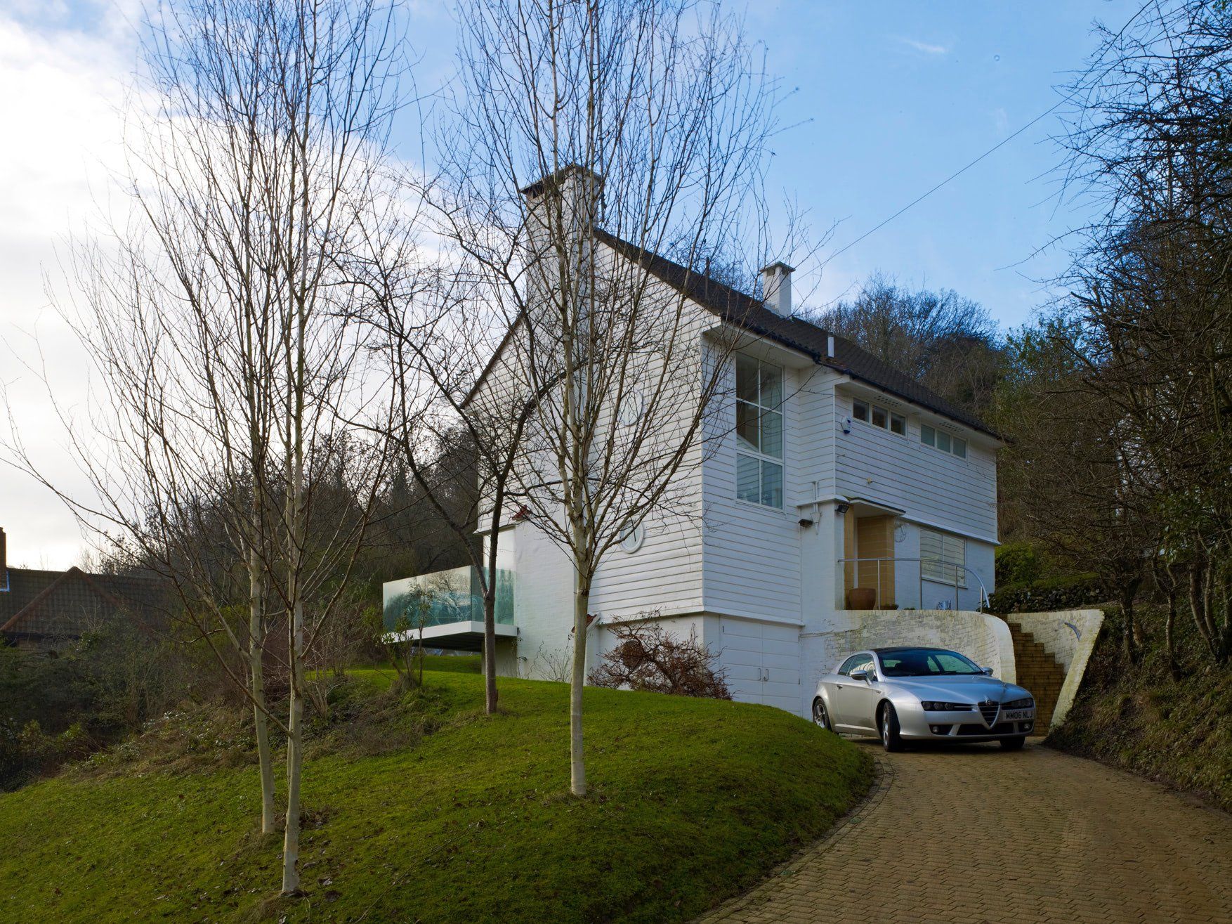 White two-story house with a silver car parked on a gravel driveway beside leafless trees and a grassy lawn.