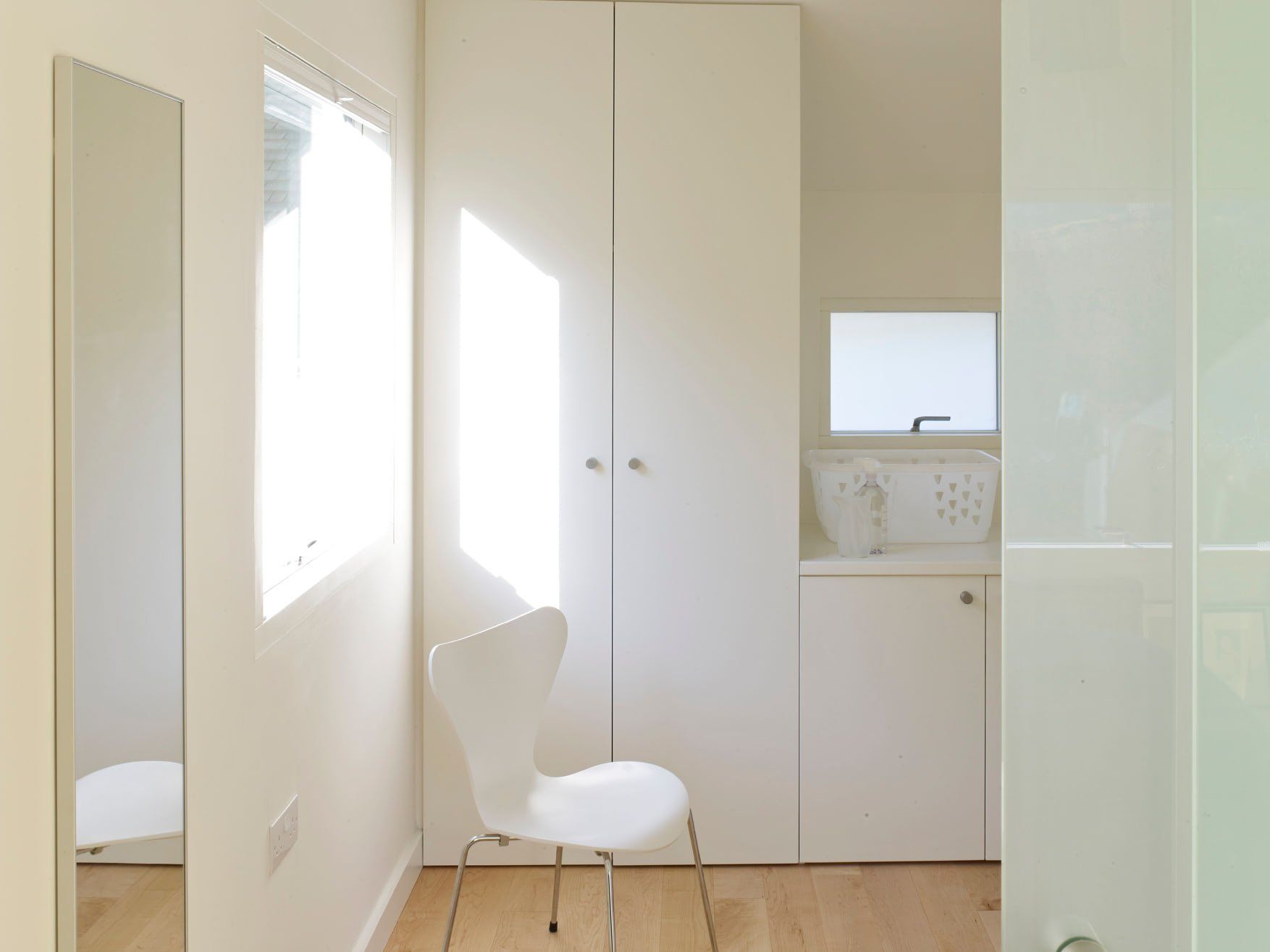 Minimal white bedroom with a chair, window light, mirrored wardrobe, and small desk nook.