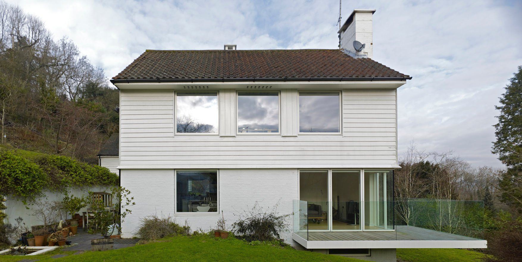 Two-story white house with a gray roof, large windows, and a small wooden deck in a leafy yard