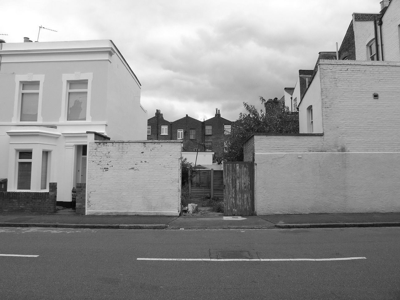 Residential street with two white houses, a walled opening, and cloudy sky.