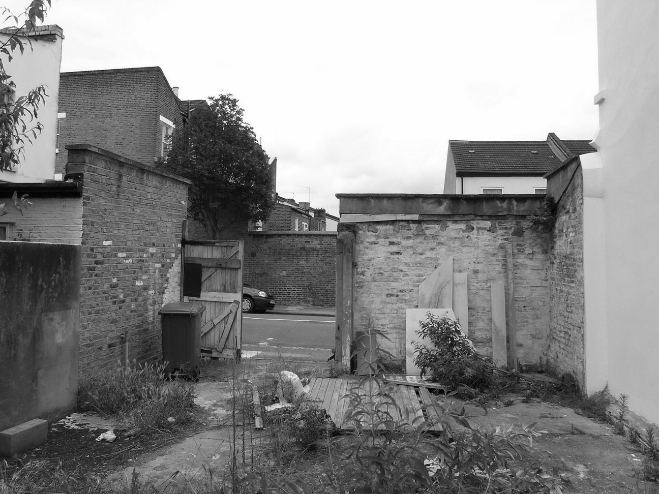 Deteriorating courtyard with old brick walls, overgrown plants, and a narrow passage under a cloudy sky