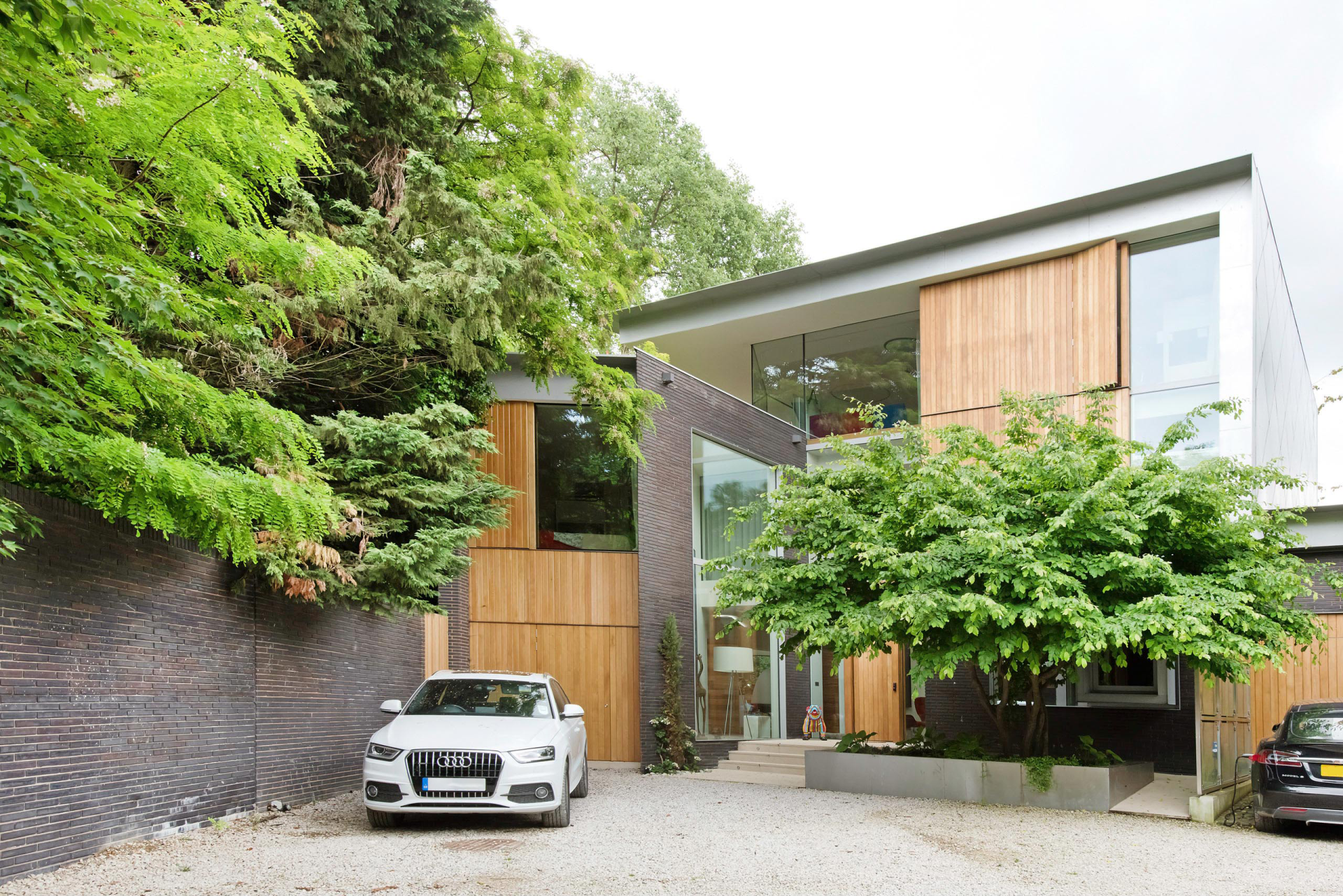 Modern two-story house with wood accents, lush trees, and a white car parked in the driveway