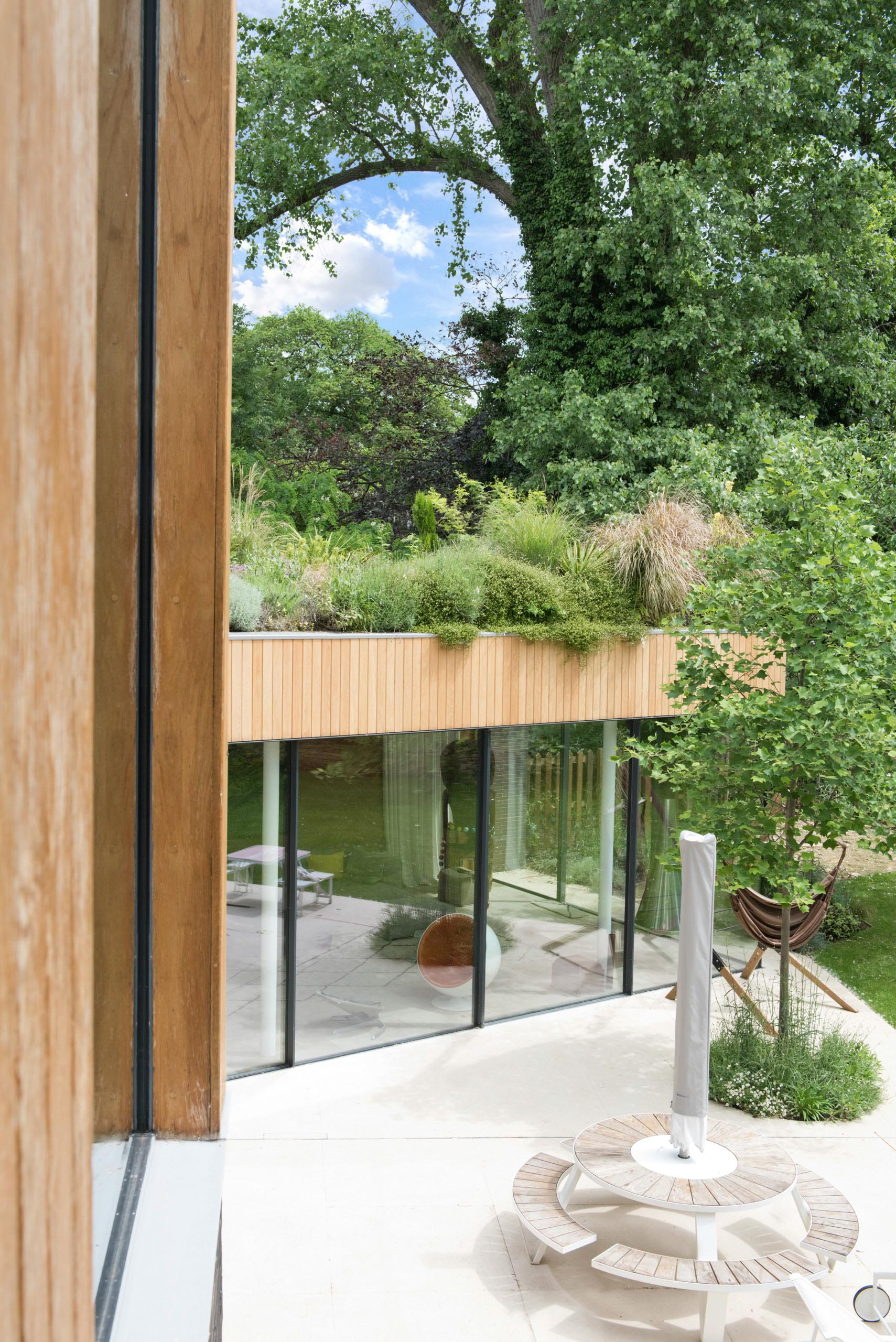 Modern house terrace with glass walls, wooden trim, and lush green trees surrounding a small patio.