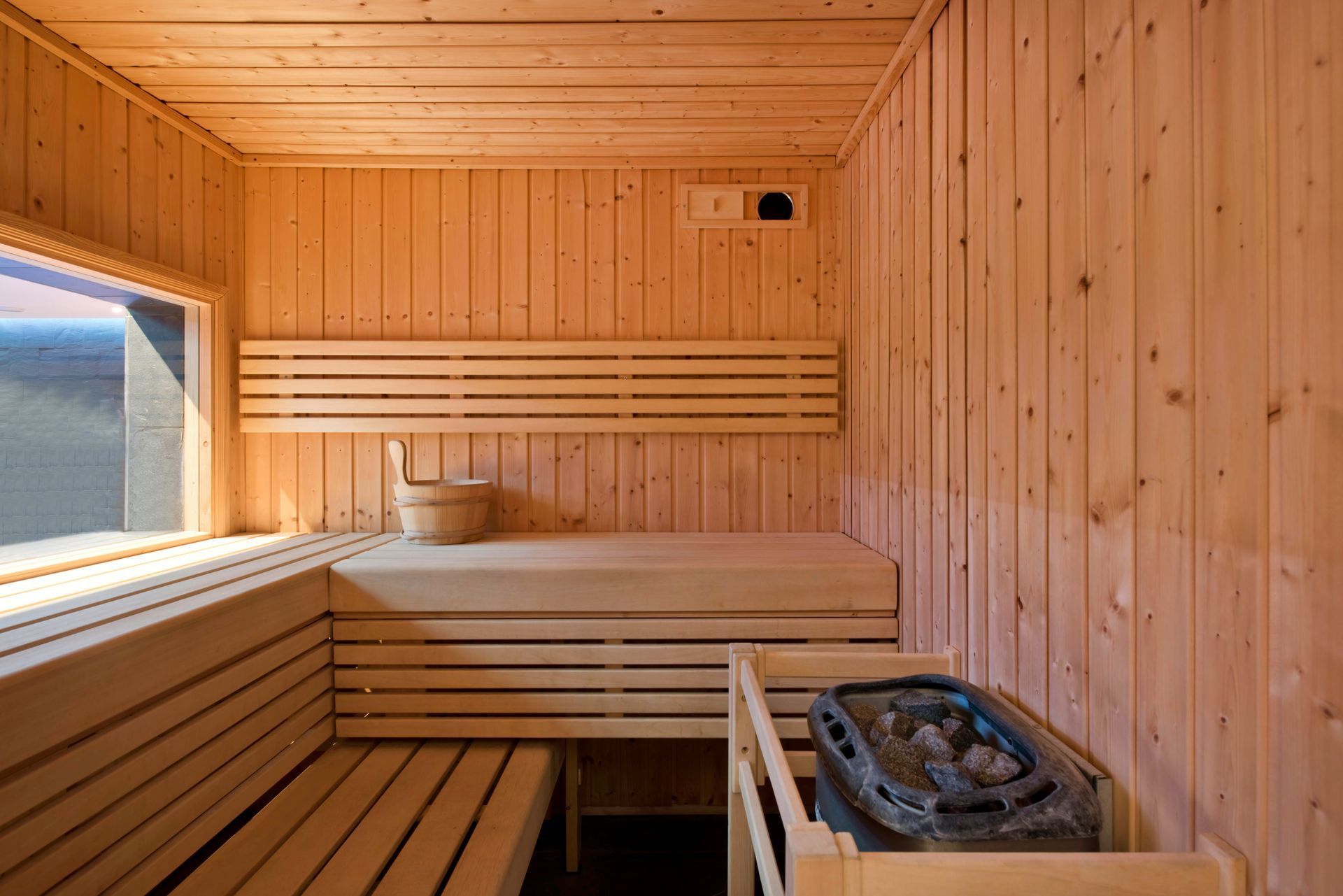 Wood-paneled sauna interior with wooden benches, heater, and a small window at left