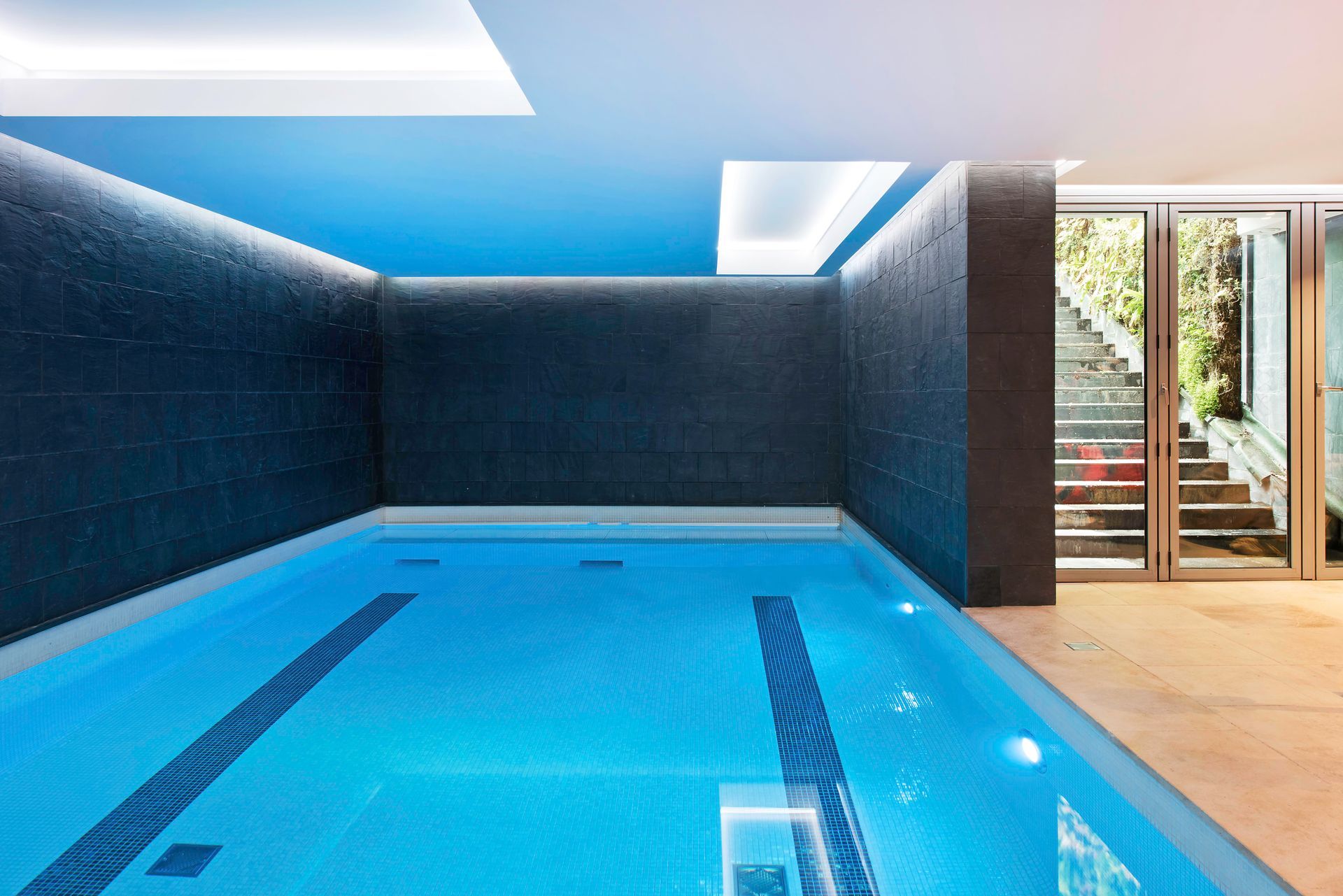 Indoor pool with blue water, dark tiled walls, and skylights beside a glass door to a patio