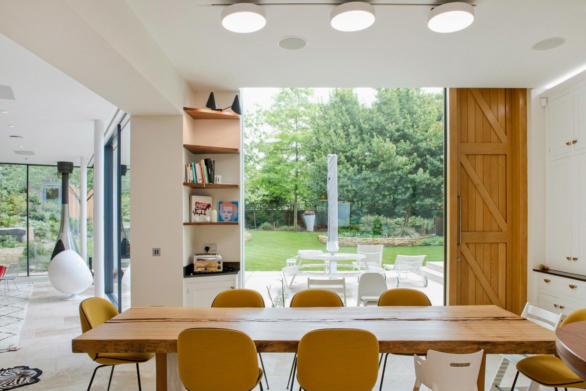 Bright modern dining room with a long wooden table, yellow chairs, and open view to a green garden.
