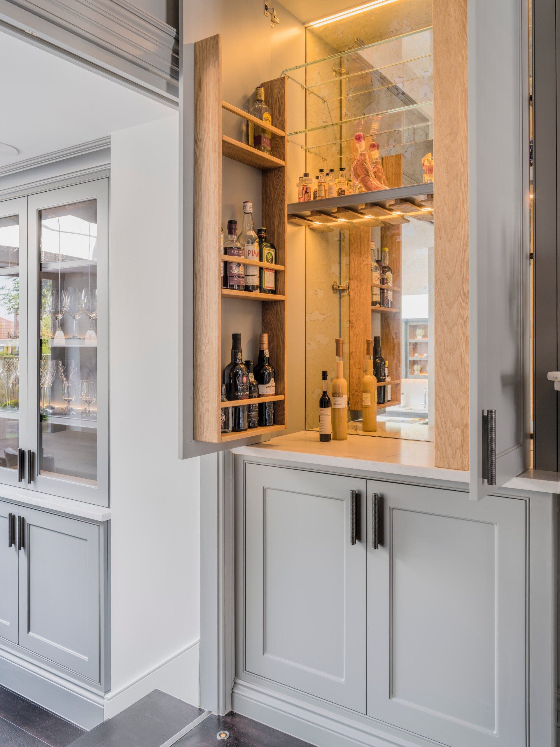 White built-in bar cabinet with glass shelves, bottles, and warm backlighting in a modern room