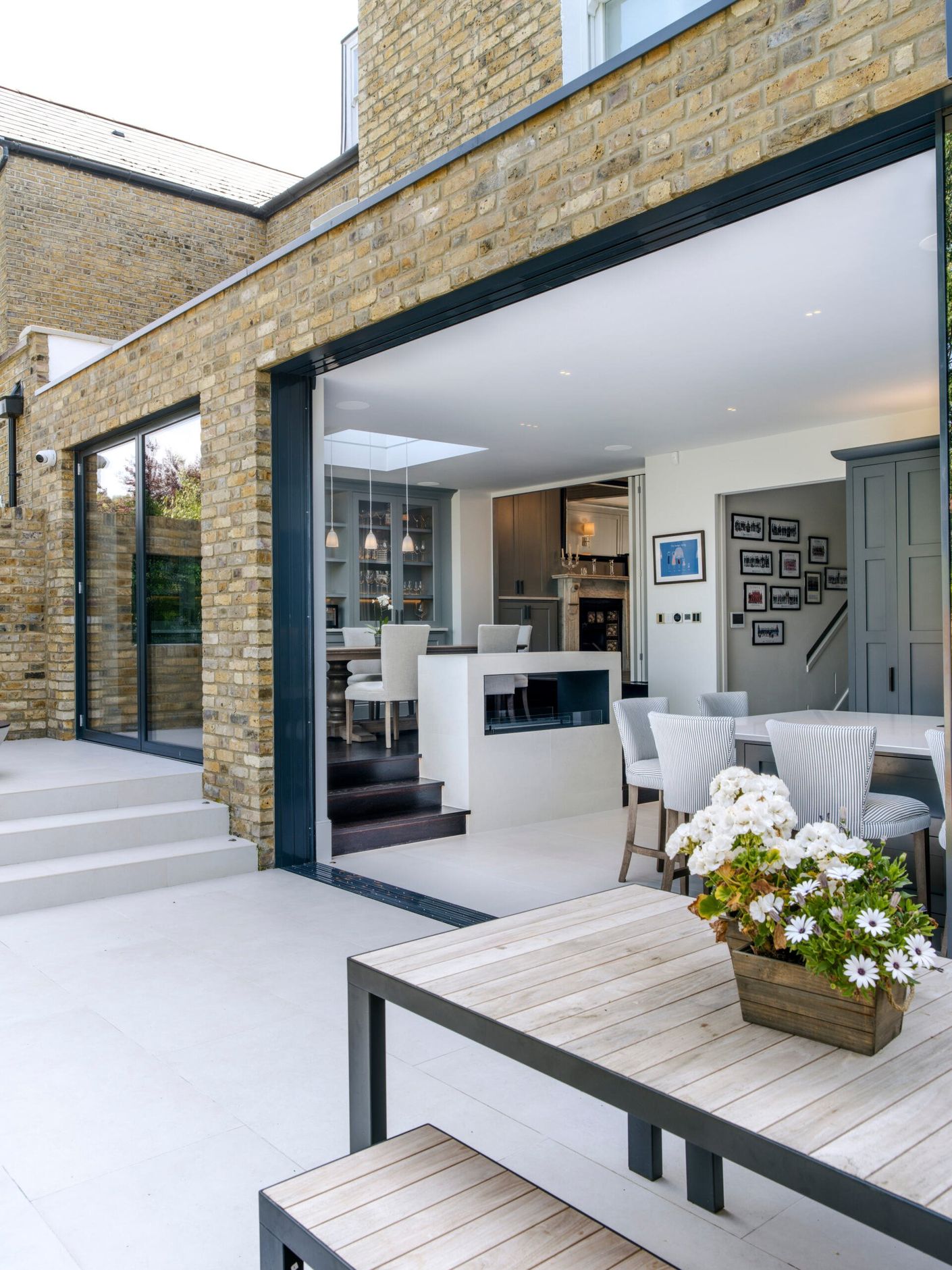 Modern patio with open sliding doors, white seating, and a potted flower arrangement outside a brick home.