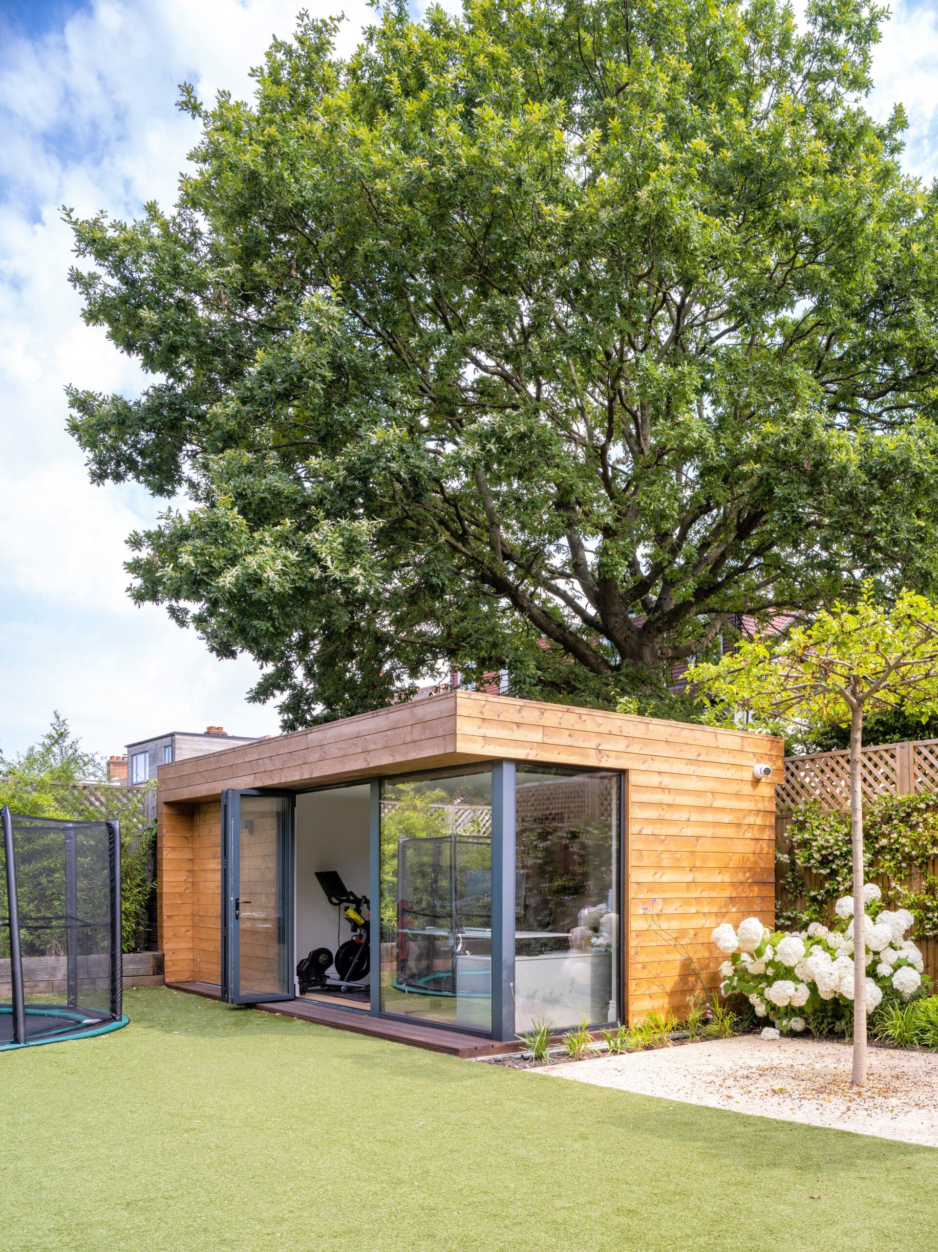 Small modern wooden garden office with glass doors beside a large tree and artificial grass lawn.