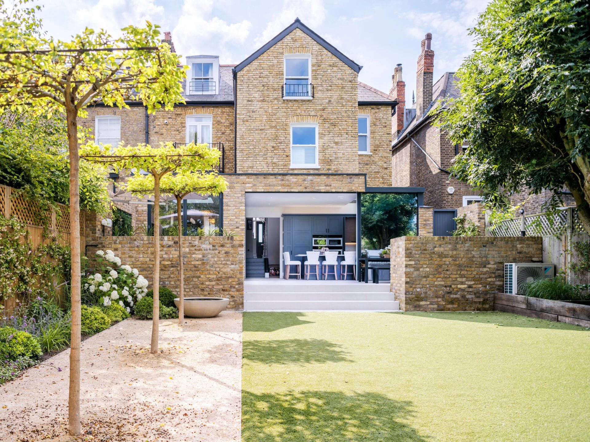 Rear garden with artificial lawn, stone patio, and a brick house with open garage-style doors