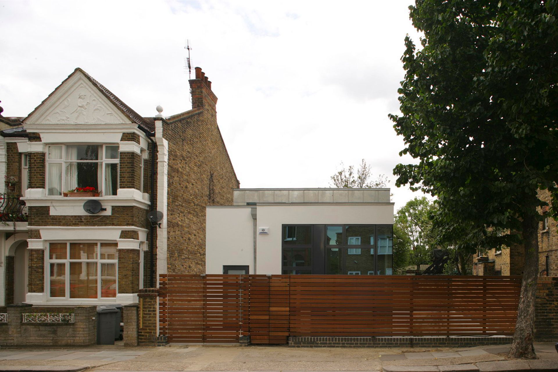 Street view of a white house beside a modern building, with a wooden fence and trees in front.
