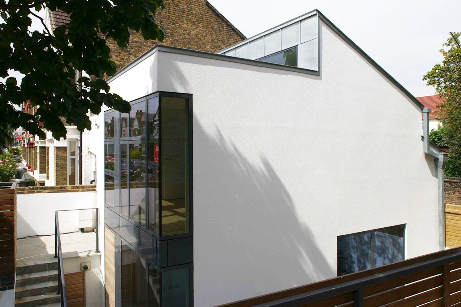 Modern white house with sloped roof and glass balcony beside trees and a wooden fence