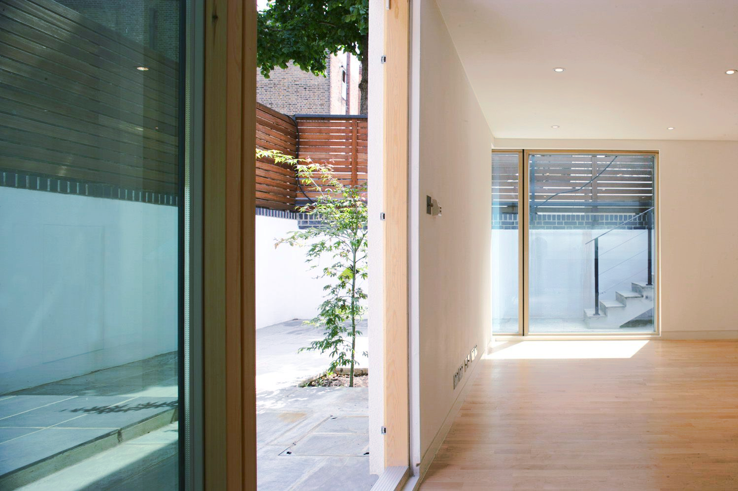 Modern hallway with wood floor, cream walls, and large windows opening to a bright courtyard.