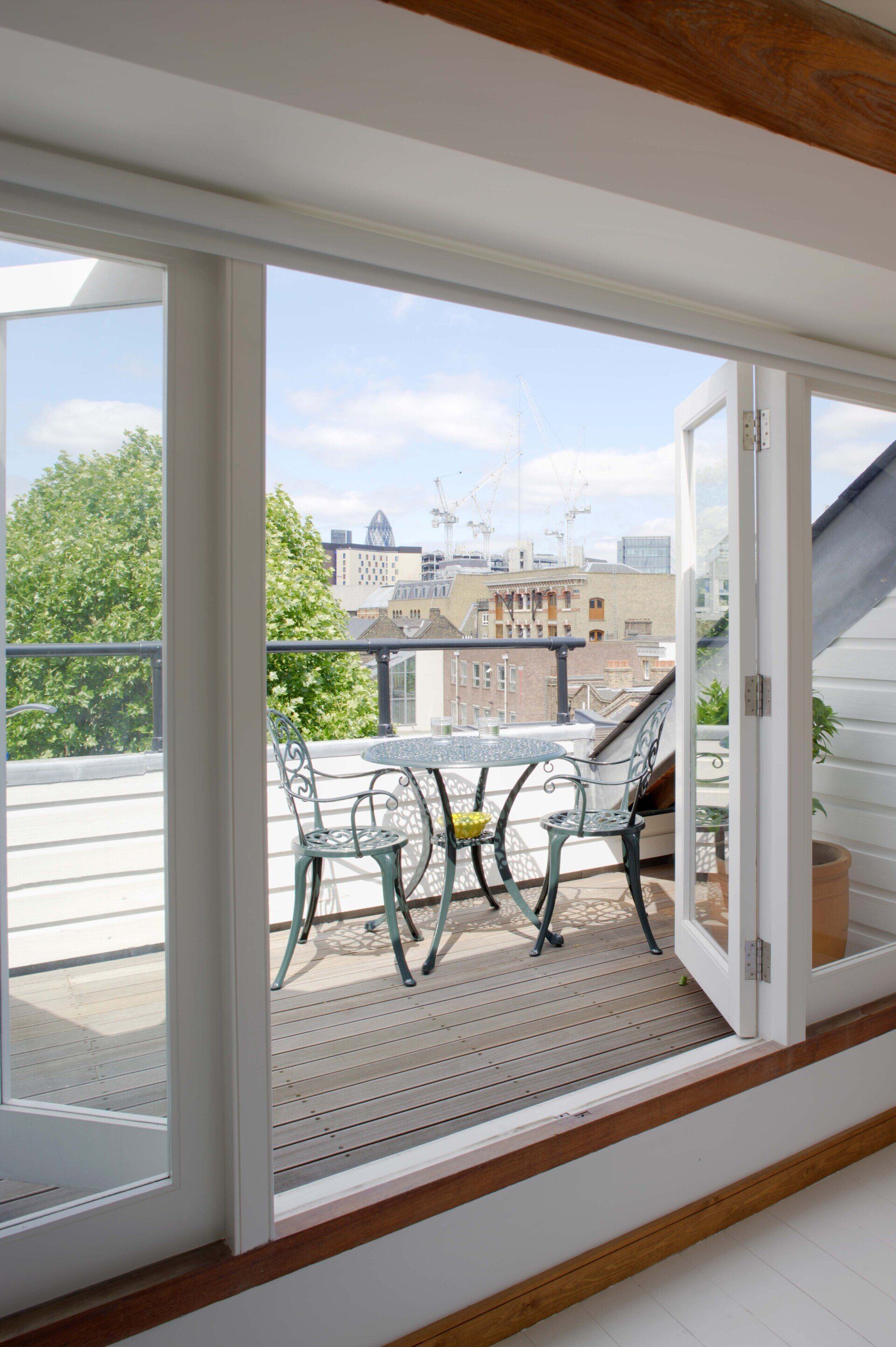 Balcony with metal table and chairs, viewed through an open window, overlooking city rooftops and trees