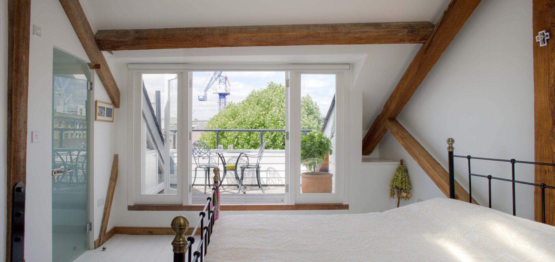 Bright attic room with exposed beams, white walls, and open French doors to a green balcony.