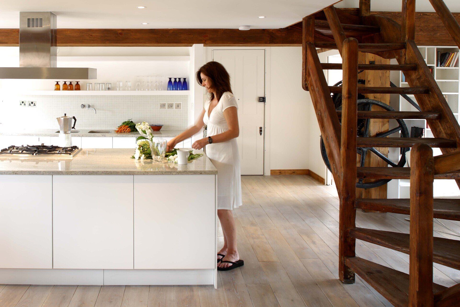 Woman in a bright modern kitchen beside a white island and wooden staircase