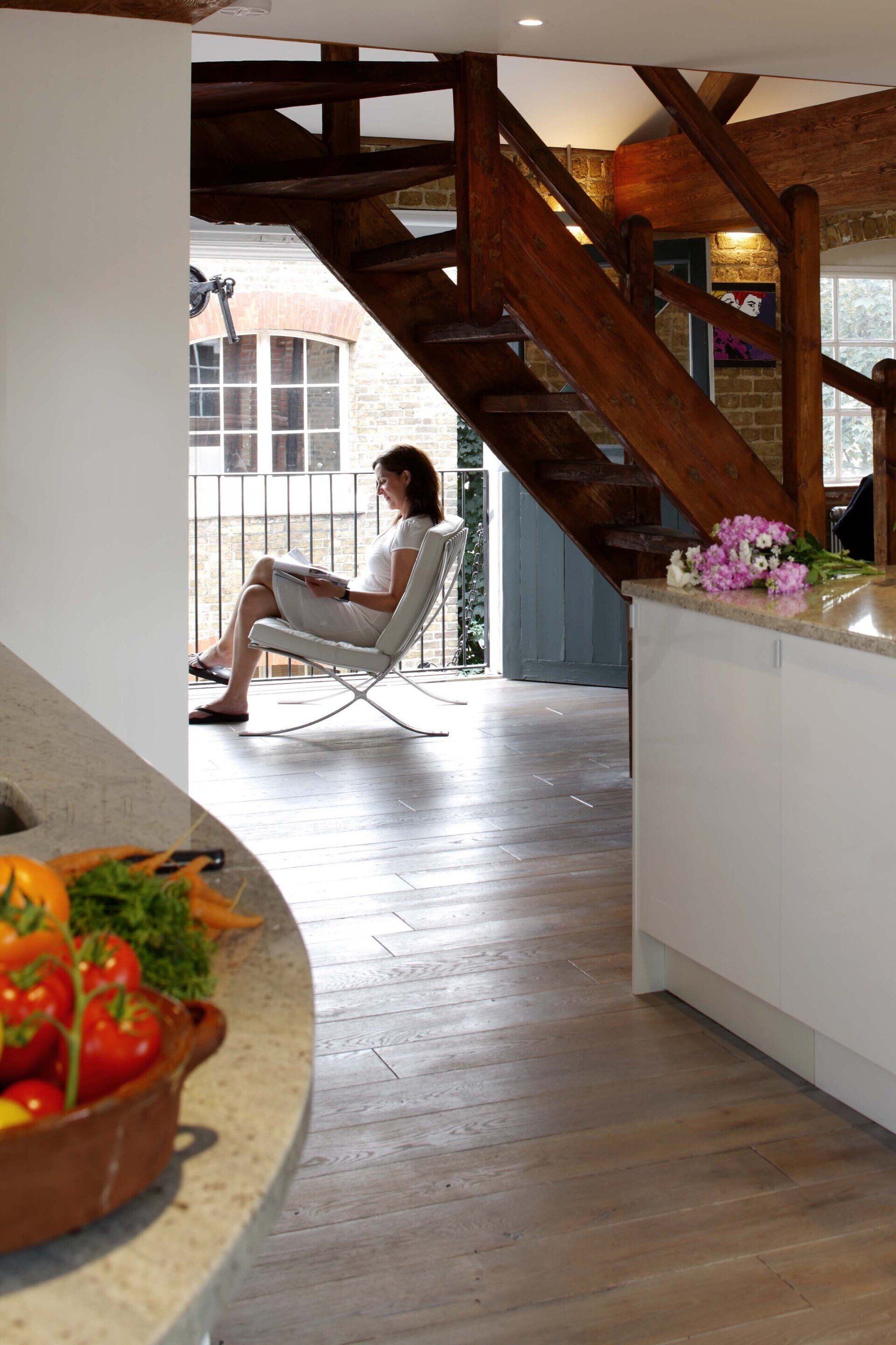 Woman relaxing in a white chair under a wooden staircase in a bright kitchen-living space