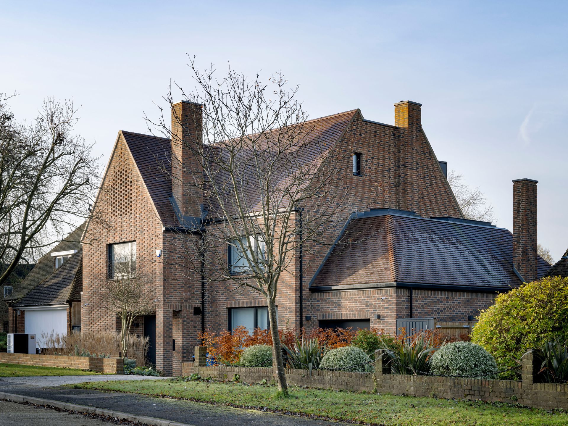 Brick house with steep roof, chimneys, and a fenced front yard in a suburban neighborhood