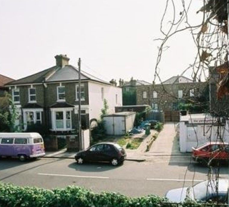 Residential street with parked cars beside brick houses and a narrow driveway under a bright sky