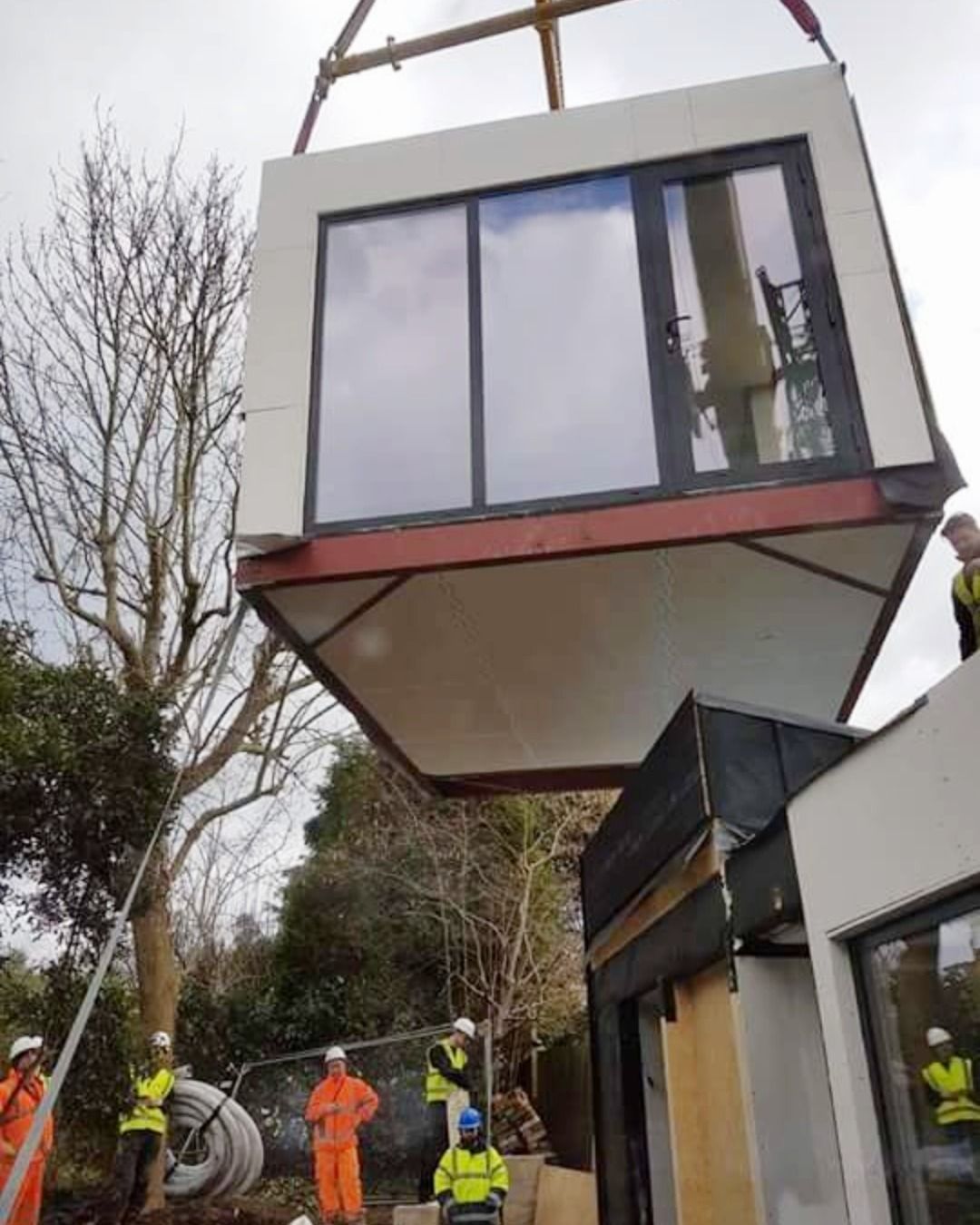 Crane lifting a modern box-shaped room into place at a construction site, with workers below
