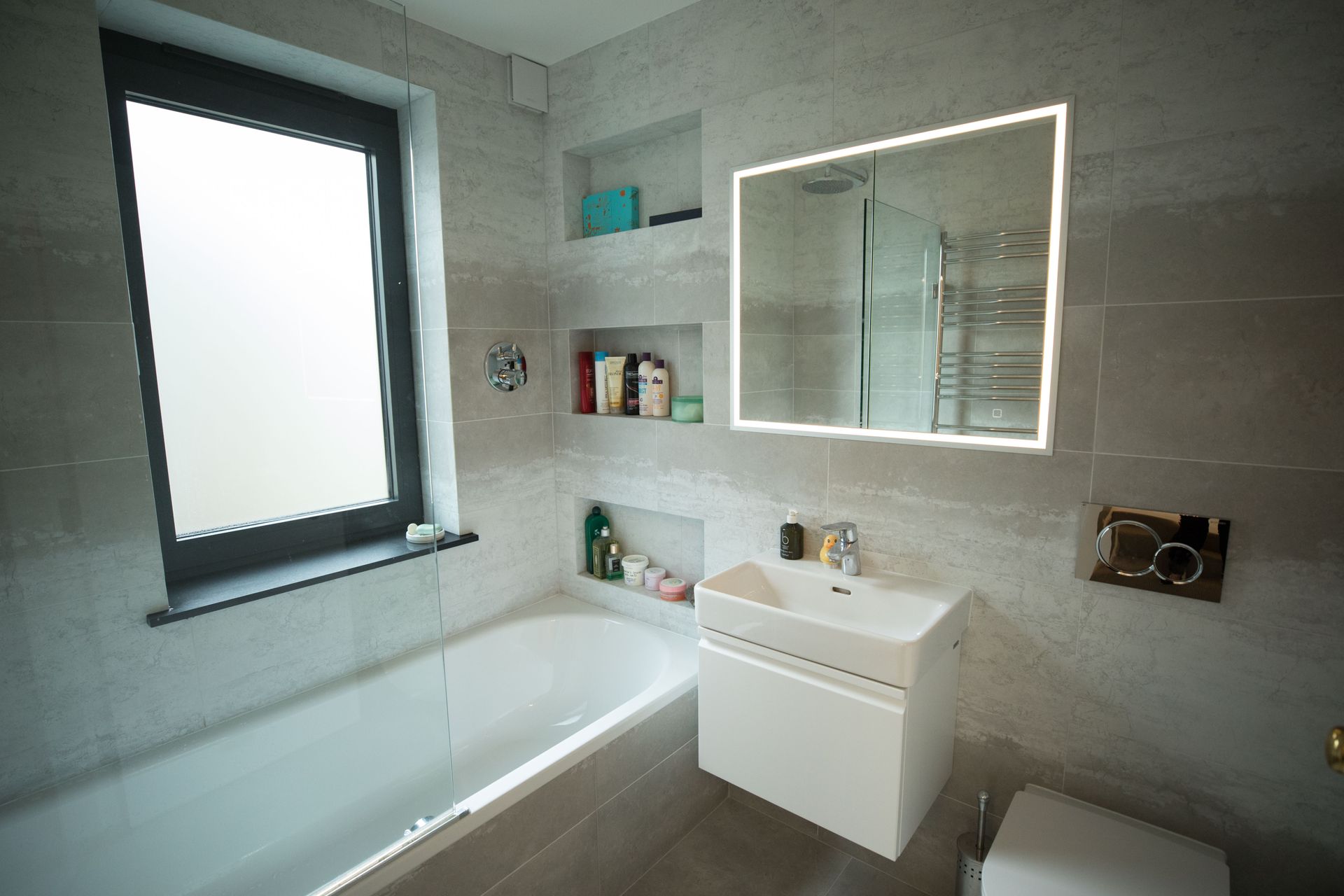 Modern gray-tiled bathroom with bathtub, vanity sink, mirror, and window