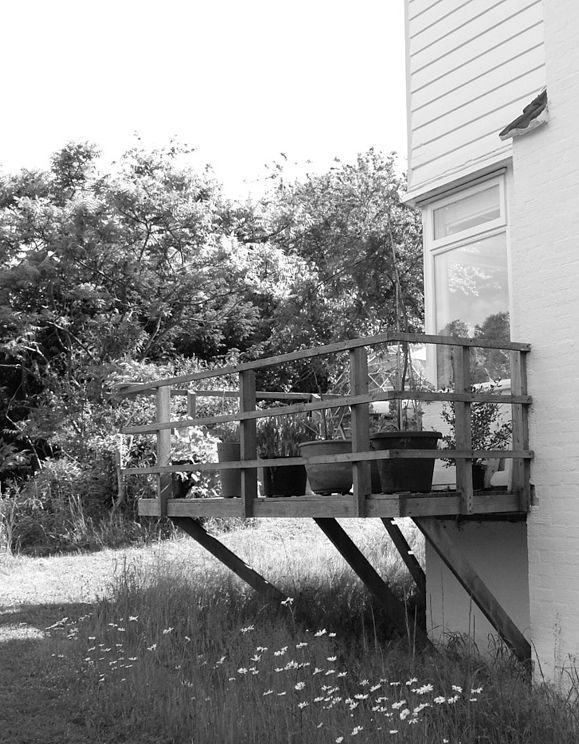 Small wooden deck on a house, raised on angled supports beside trees and grass.