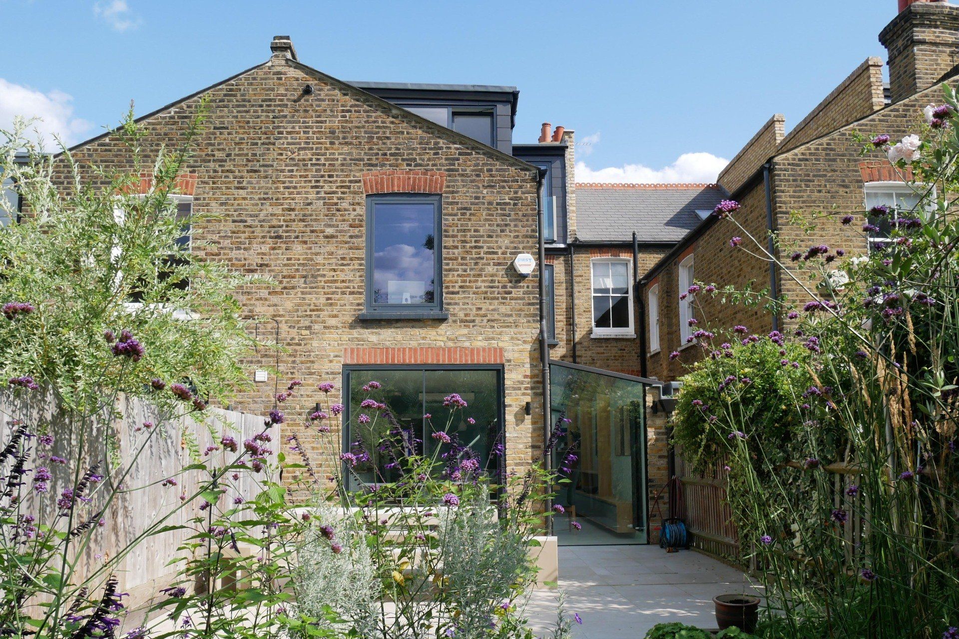 Backyard garden path between brick houses, with climbing greenery and a blue sky.