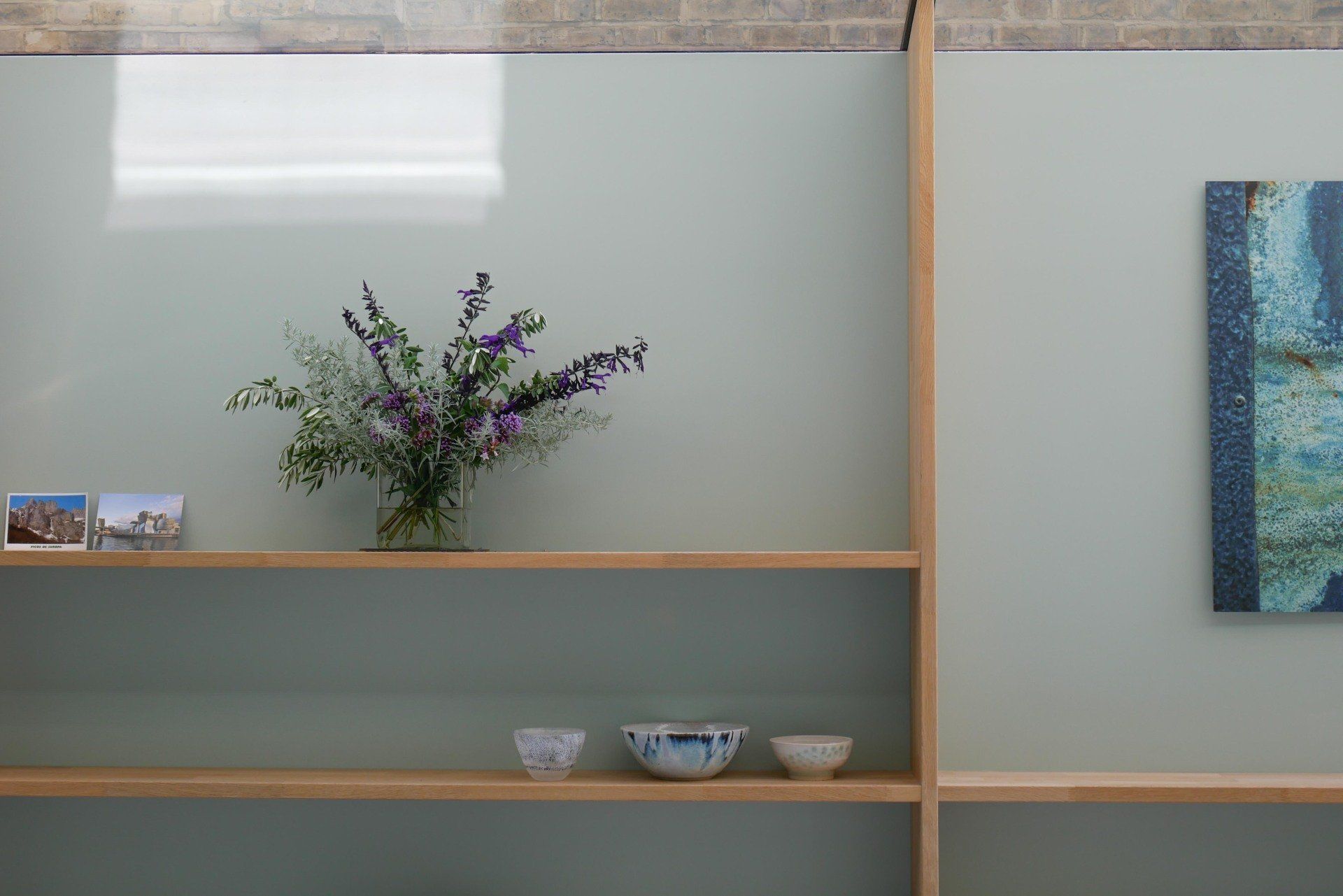 Minimalist shelf with a vase of purple flowers, small bowls, and a blue abstract painting on a pale wall
