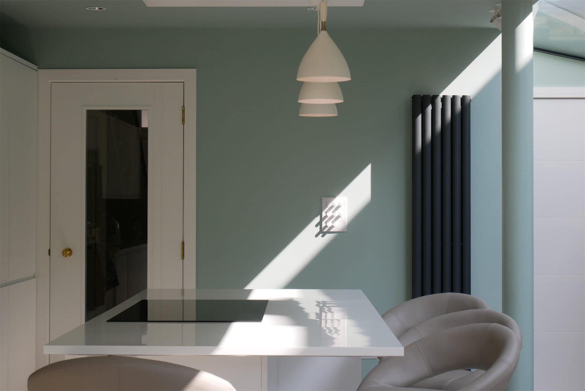 Modern dining room with mint walls, glass table, white chairs, pendant light, and sunlight casting shadows.