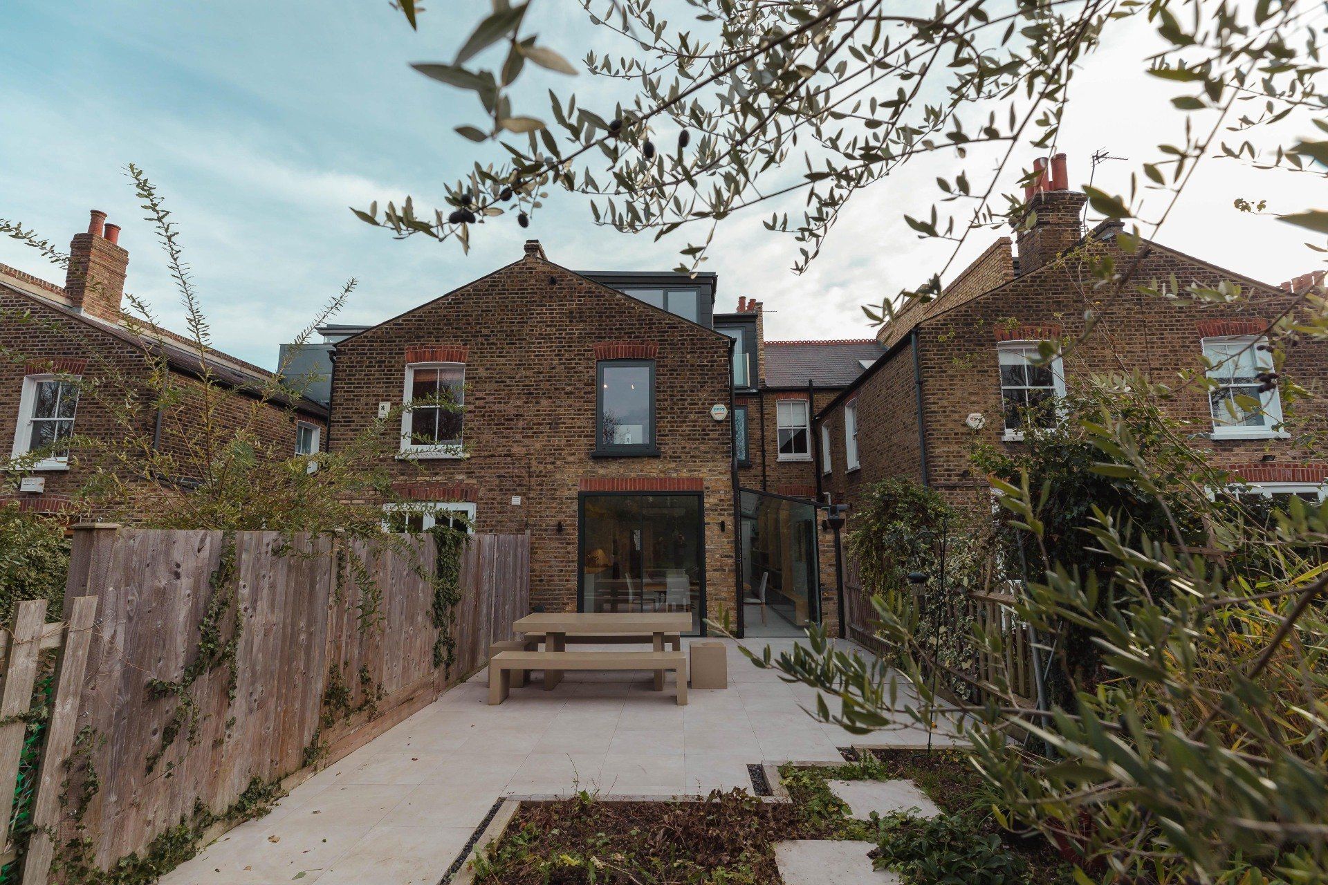 Brick house garden with patio, bench, and table, framed by trees and shrubs