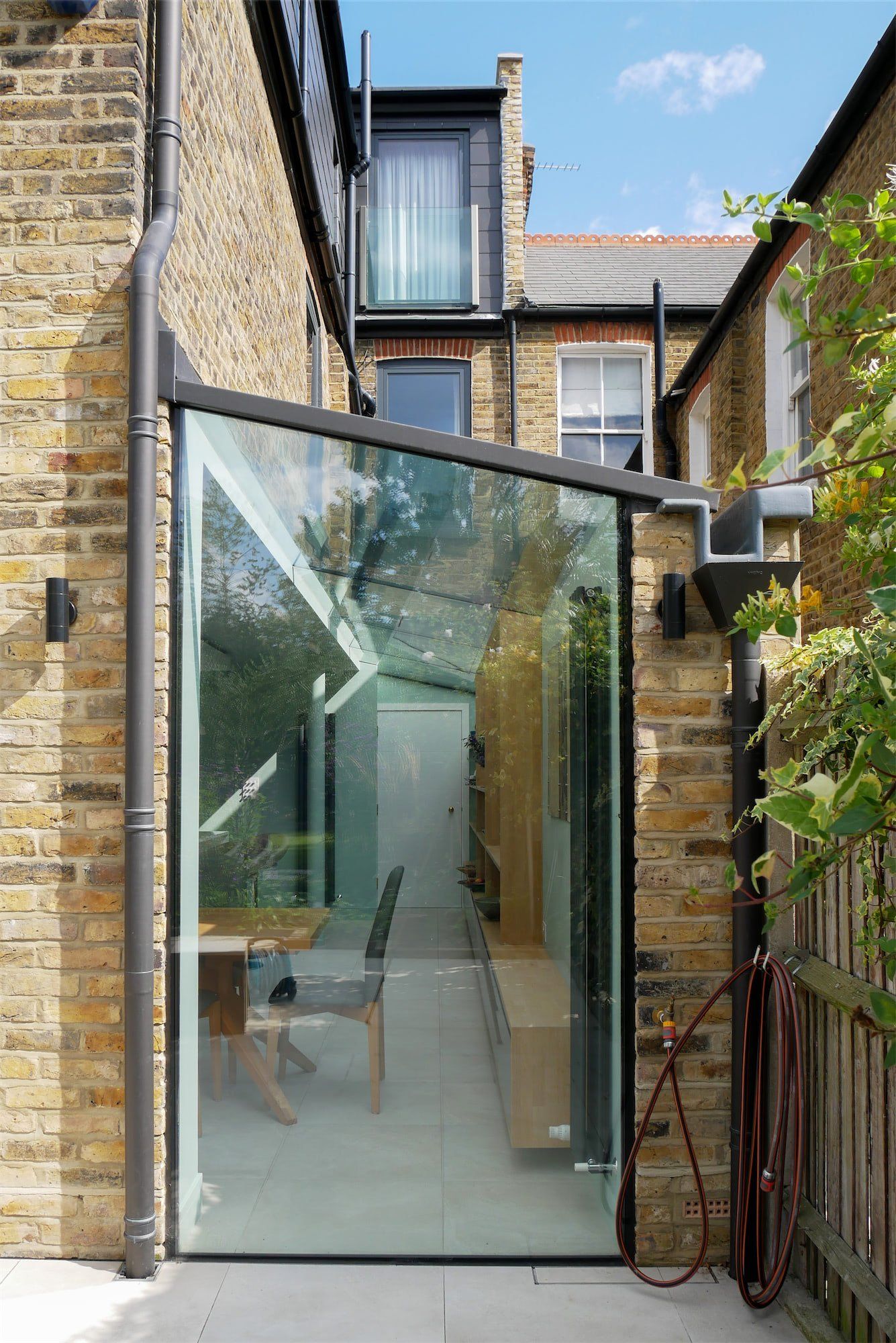 Glass extension in a narrow brick courtyard between townhouses, with a reflective slanted roof.