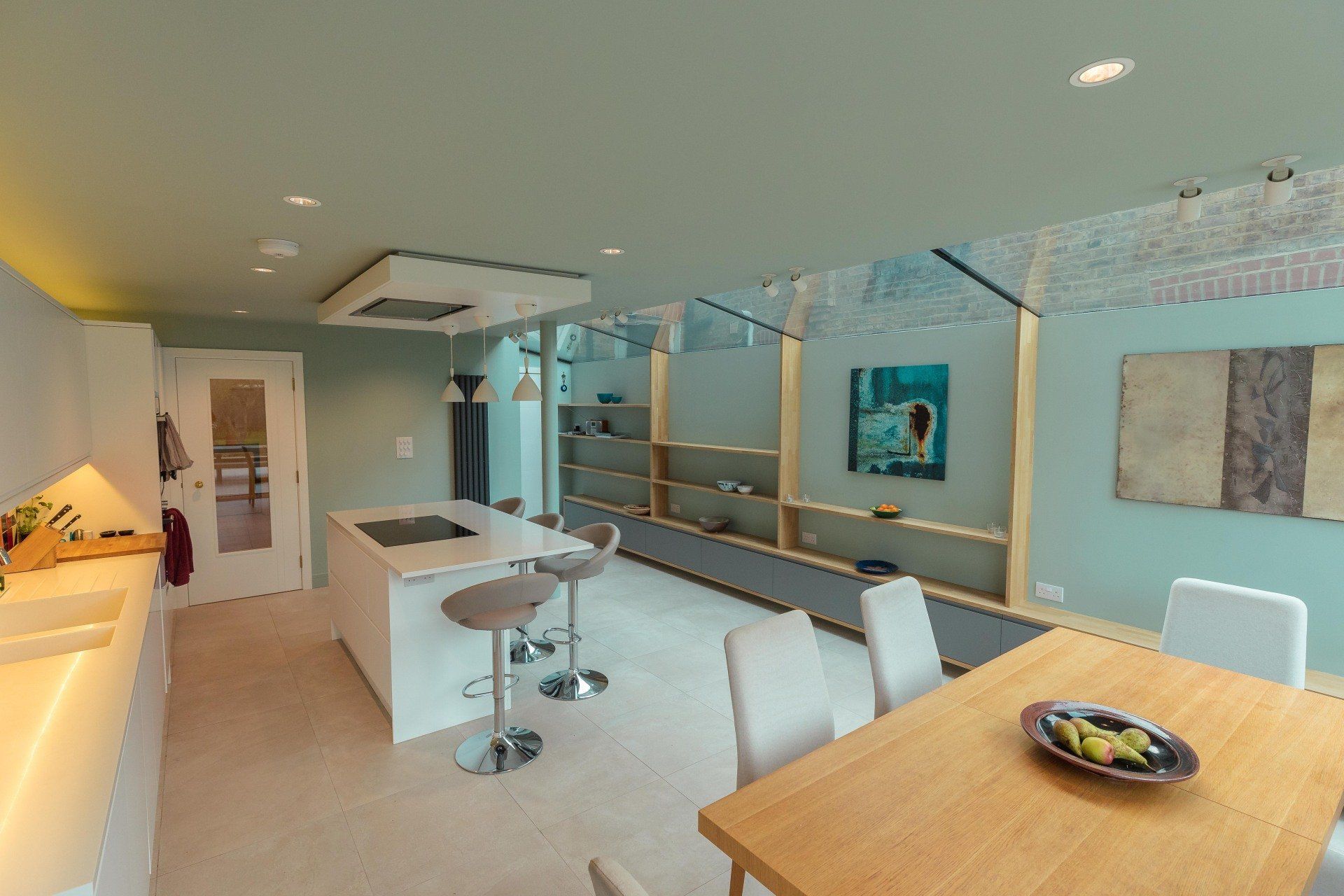 Modern open-plan kitchen and dining area with white cabinets, wood table, stools, and glass partition