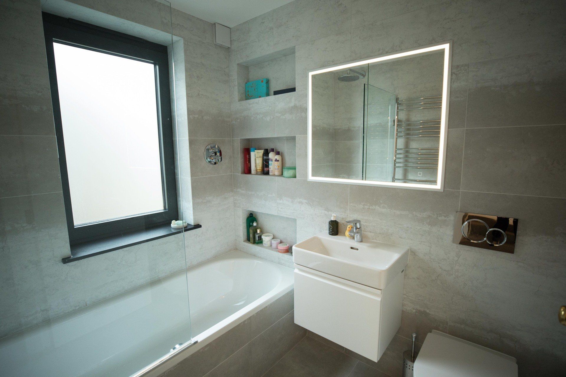 Modern gray bathroom with bathtub, window, vanity sink, mirror, and built-in shelves