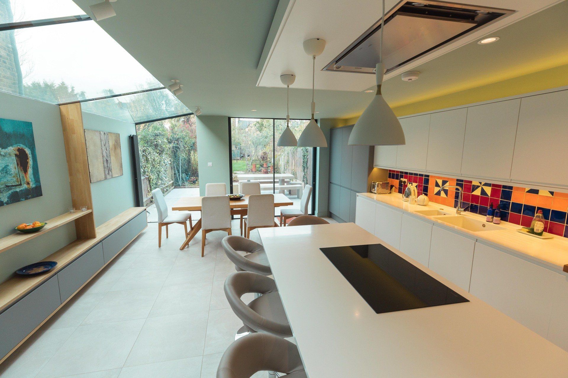 Modern minimalist kitchen with white counters, colorful backsplash, and large windows overlooking greenery