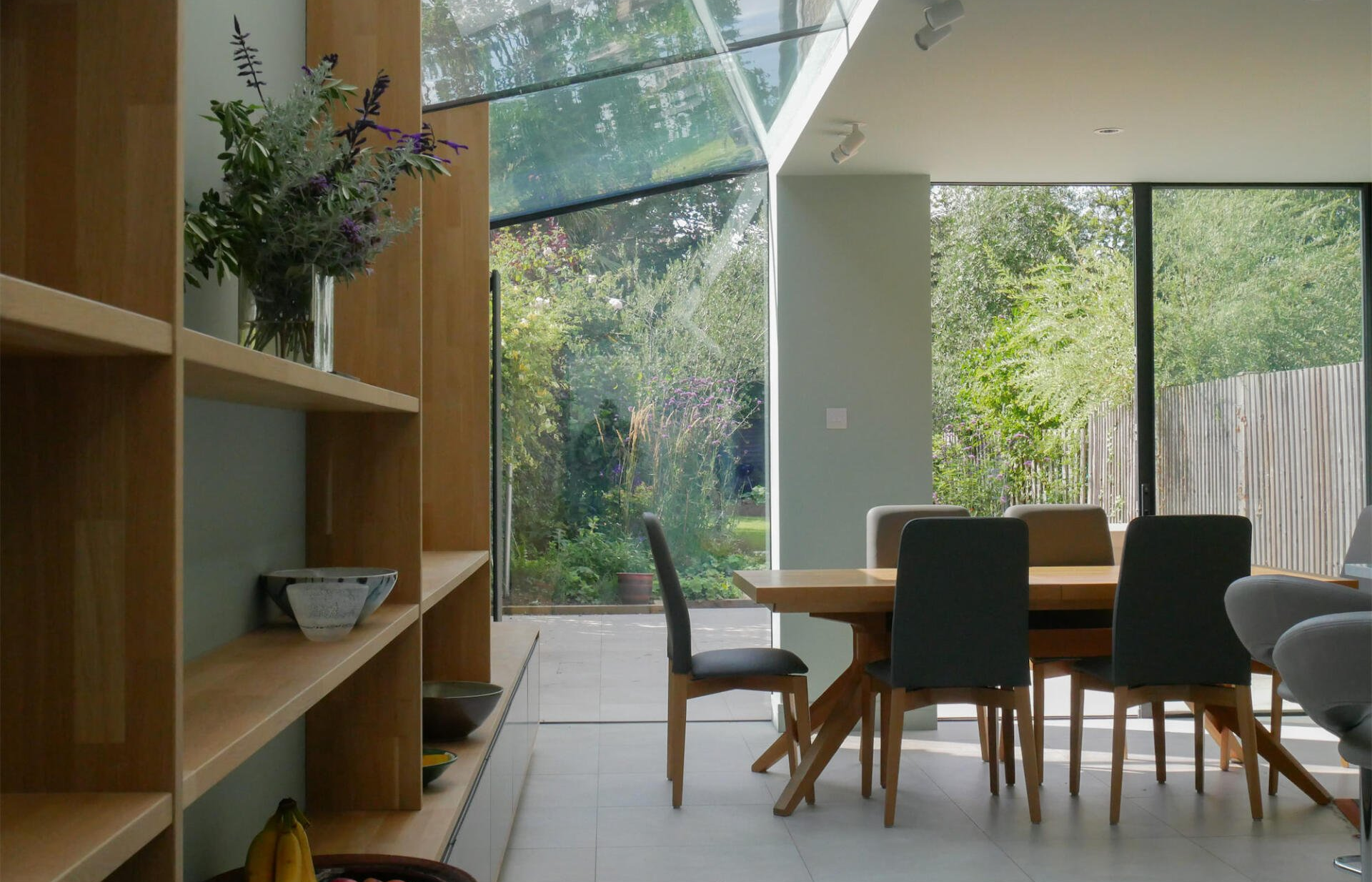 Modern dining room with large glass walls, wooden table, chairs, and sunlight overlooking a green garden
