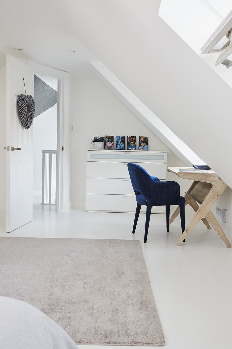 Bright white loft bedroom with desk, blue chair, dresser, and skylight under slanted ceiling