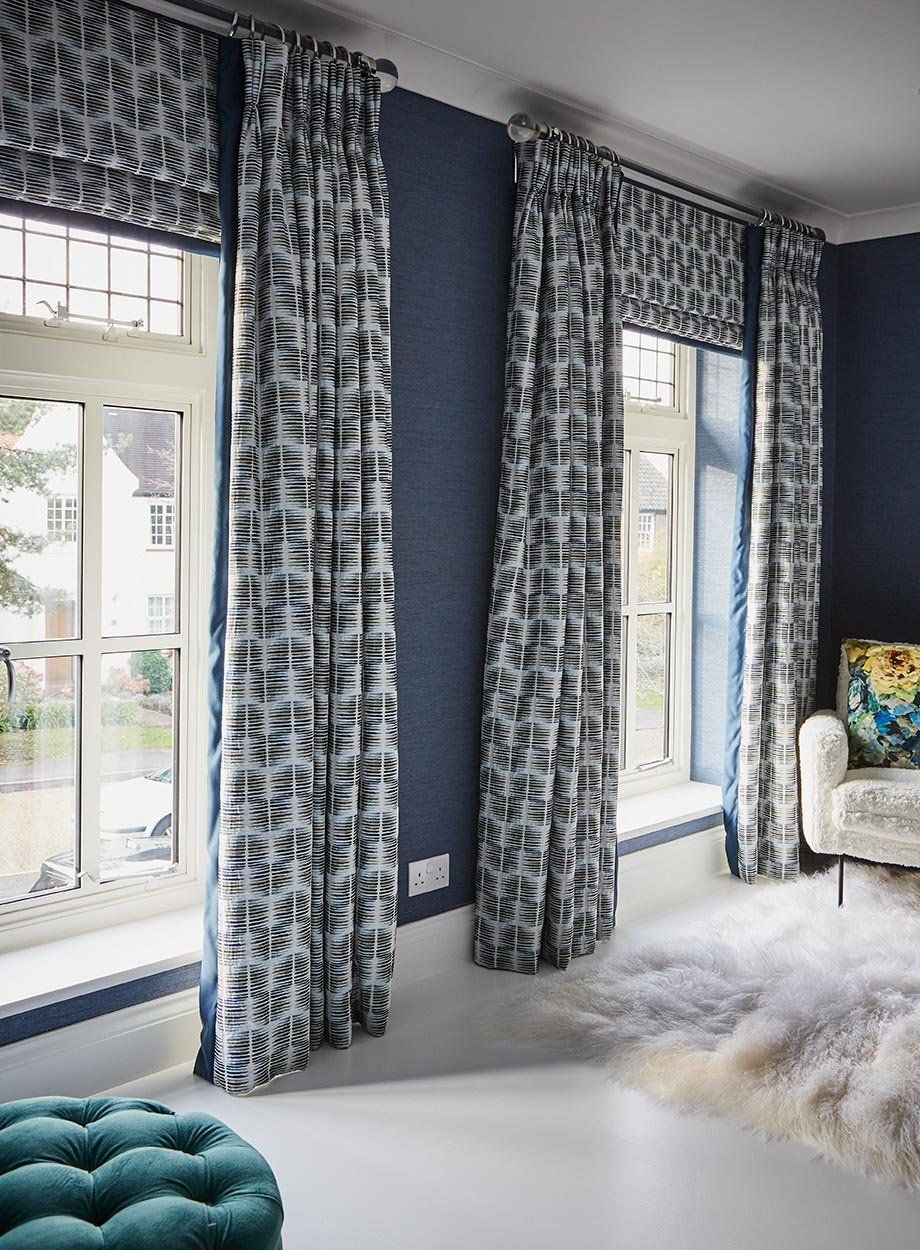 Blue patterned curtains framing tall windows in a bright, modern living room with a teal ottoman and white rug