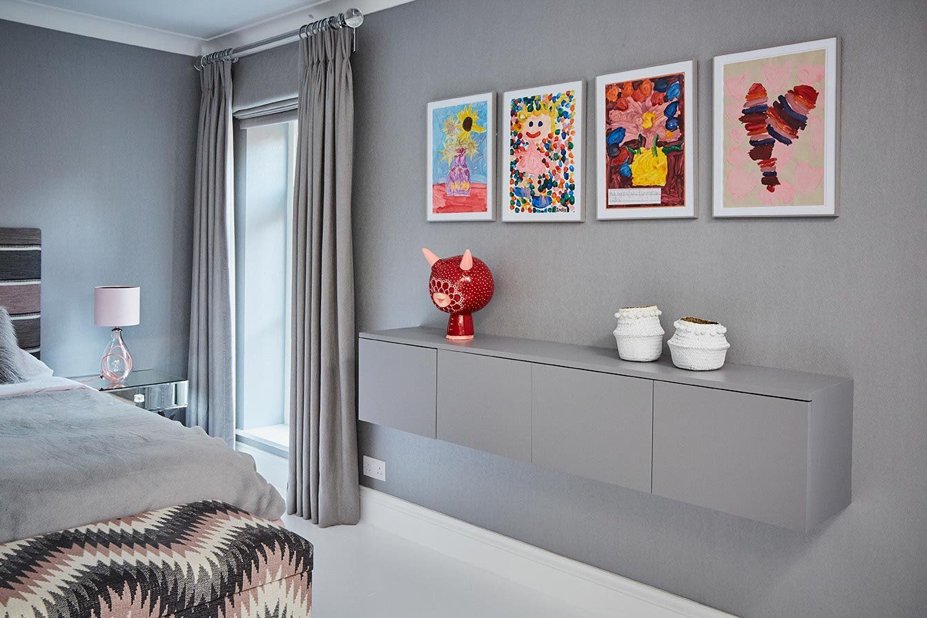 Modern bedroom with gray walls, colorful framed art, floating shelf, red vase, and bed near a window