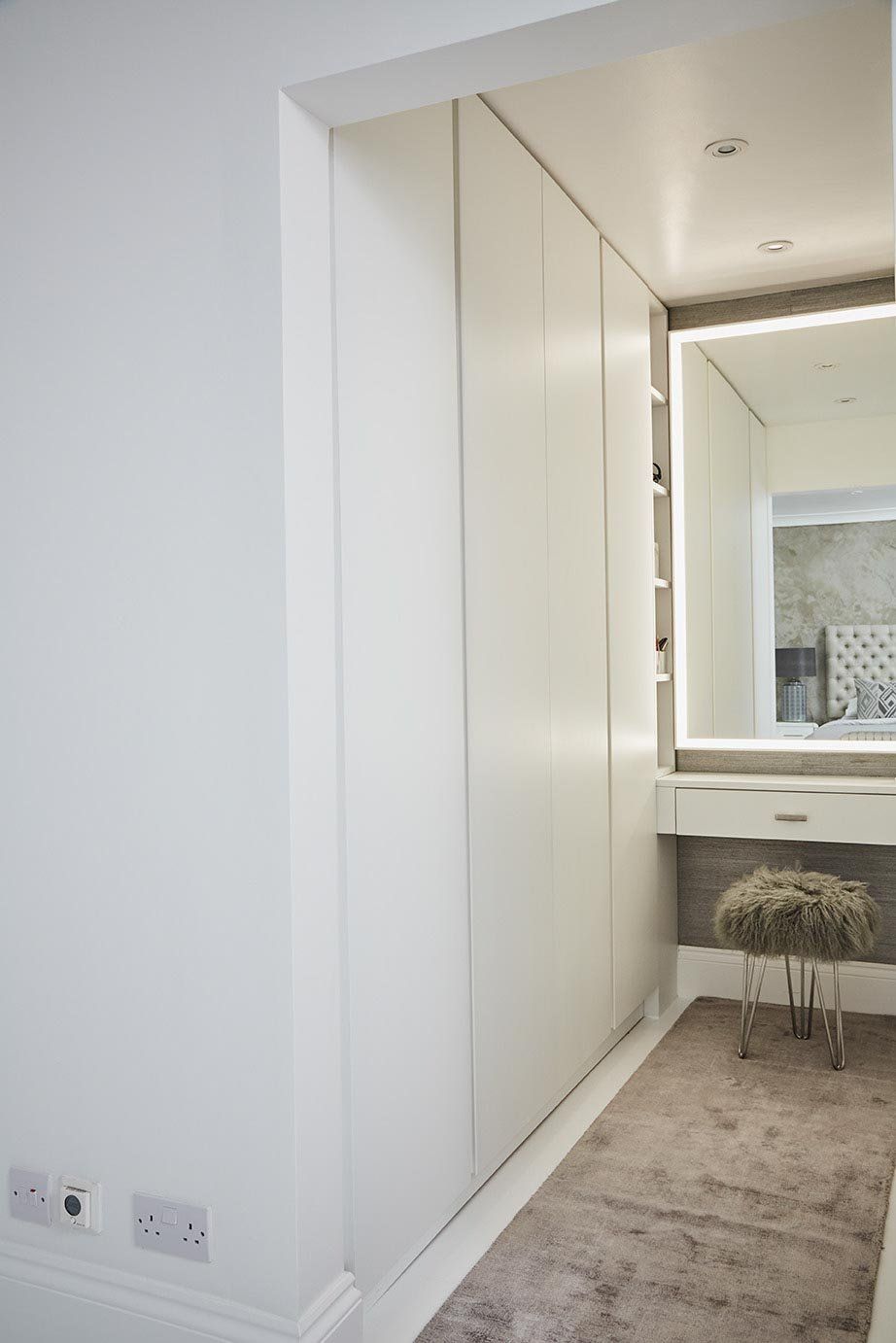 Modern white hallway with mirrored closet doors, vanity light, and a small stool on a rug.