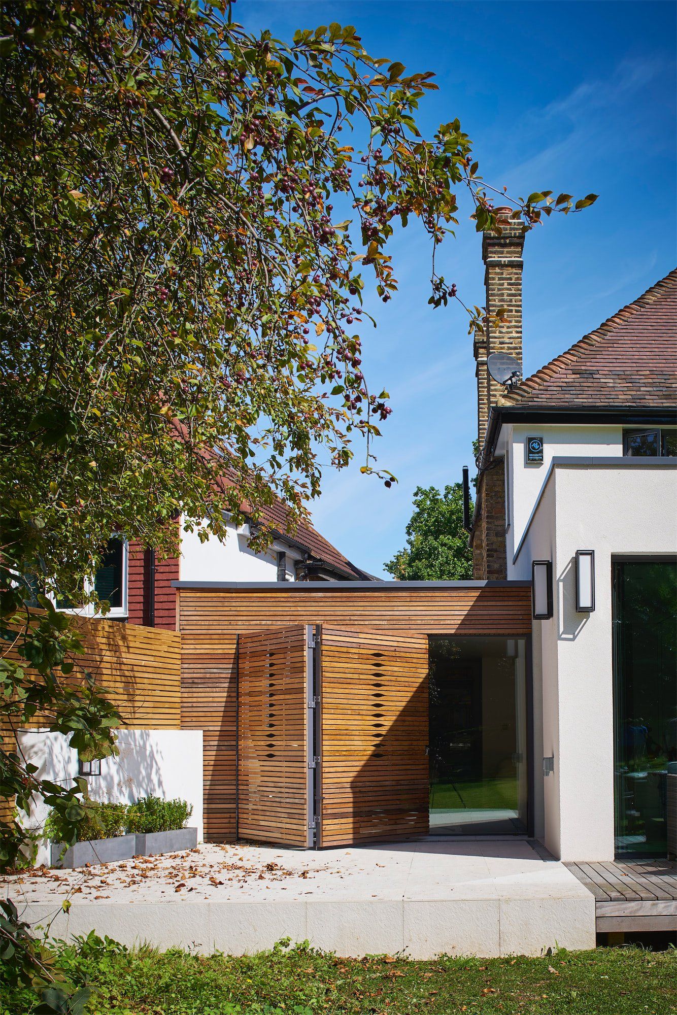 Modern house entrance with wooden gate, white walls, and a tree under a blue sky