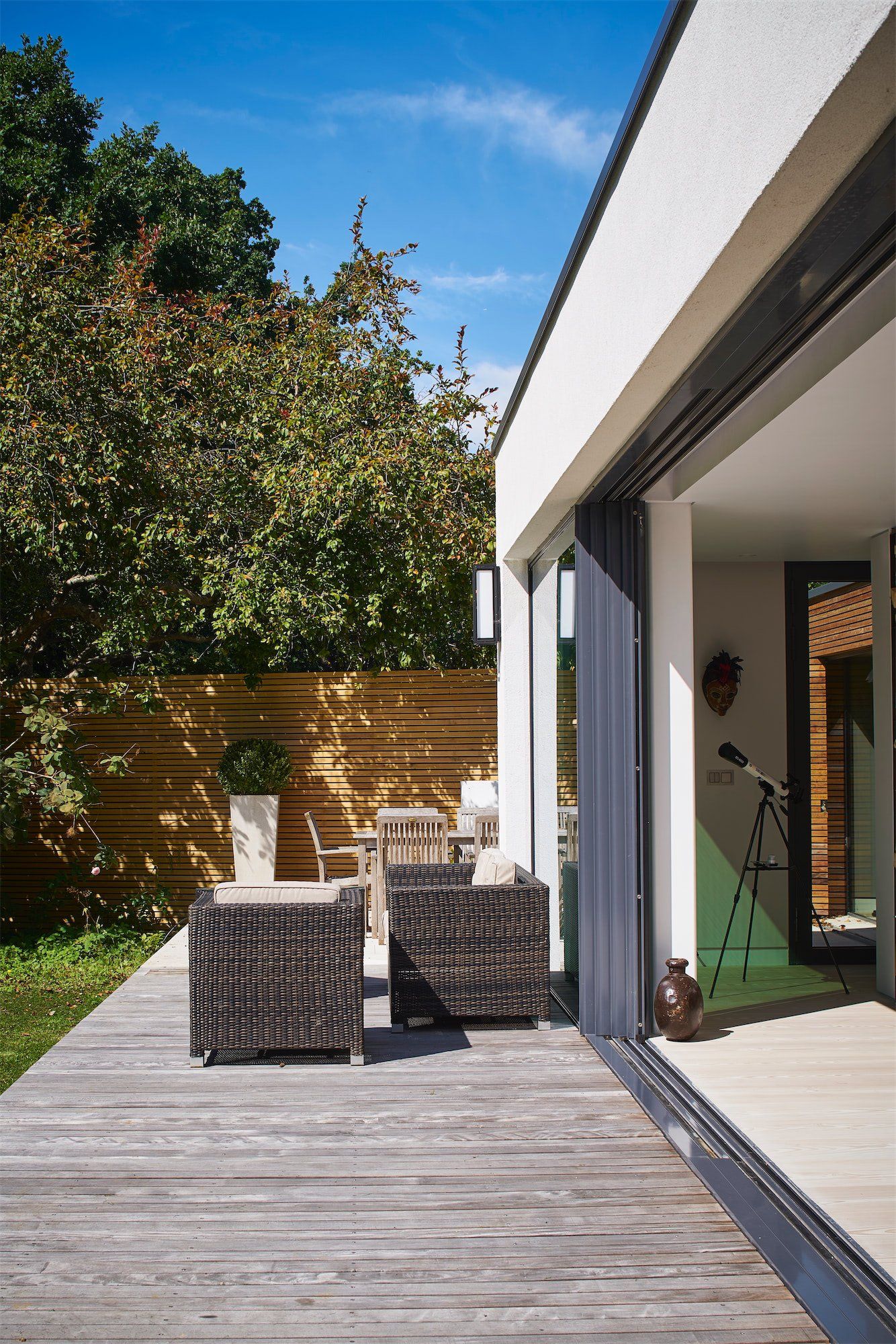 Modern patio with wicker chairs, wooden deck, and sliding glass doors beside a garden wall
