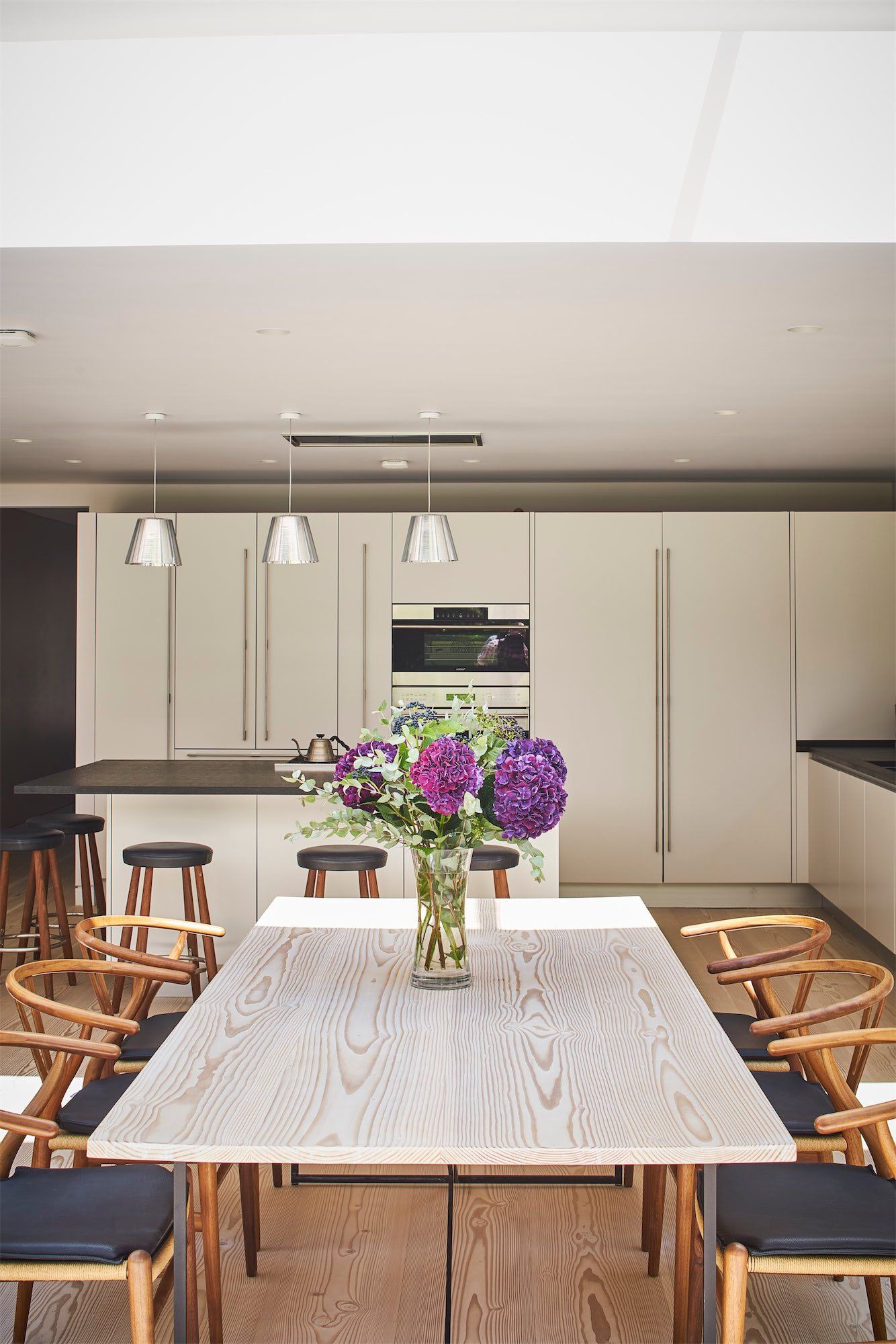 Modern kitchen with long dining table, wooden chairs, and a vase of purple flowers