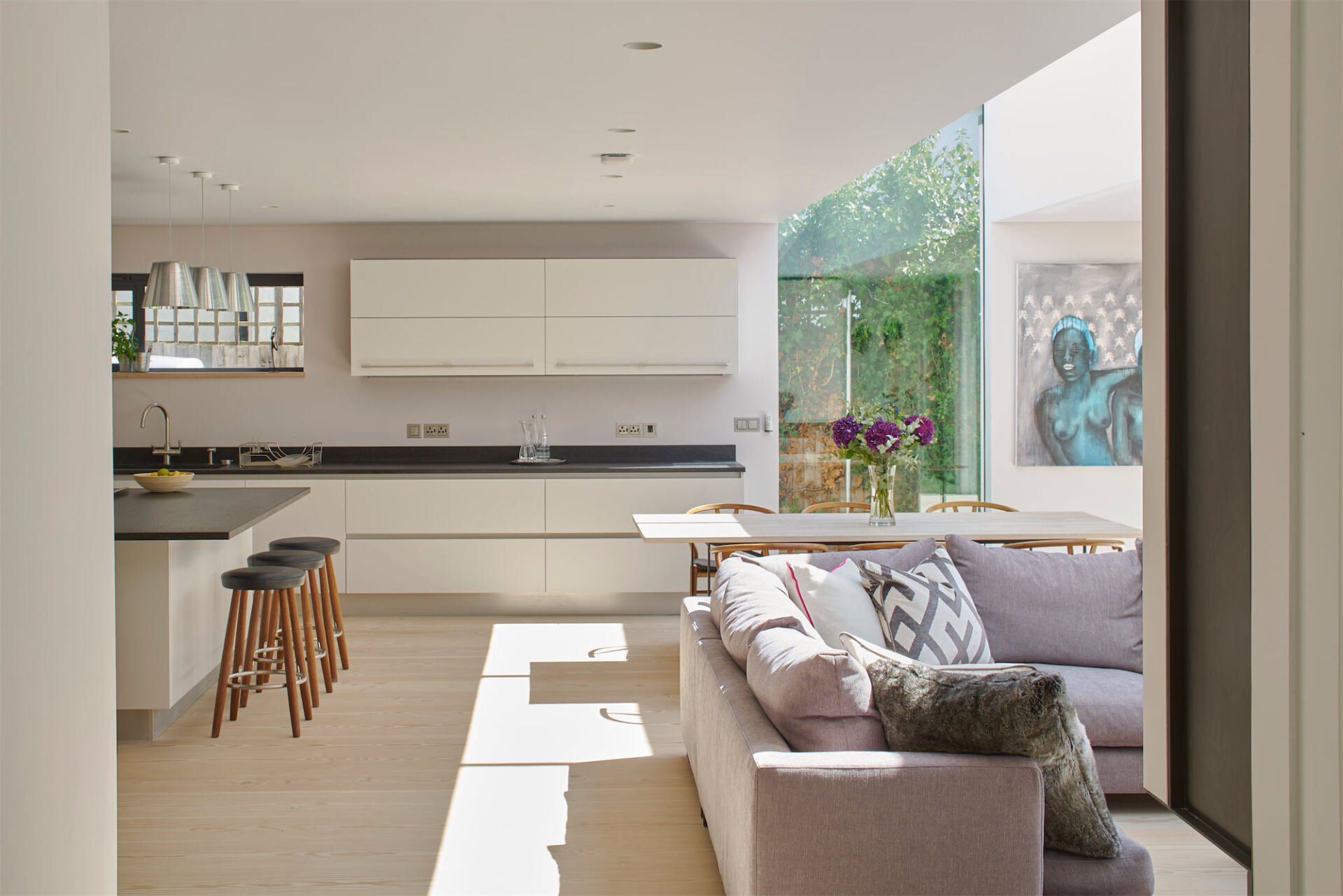 Bright modern living room and kitchen with white walls, gray sofa, wooden stools, and garden view