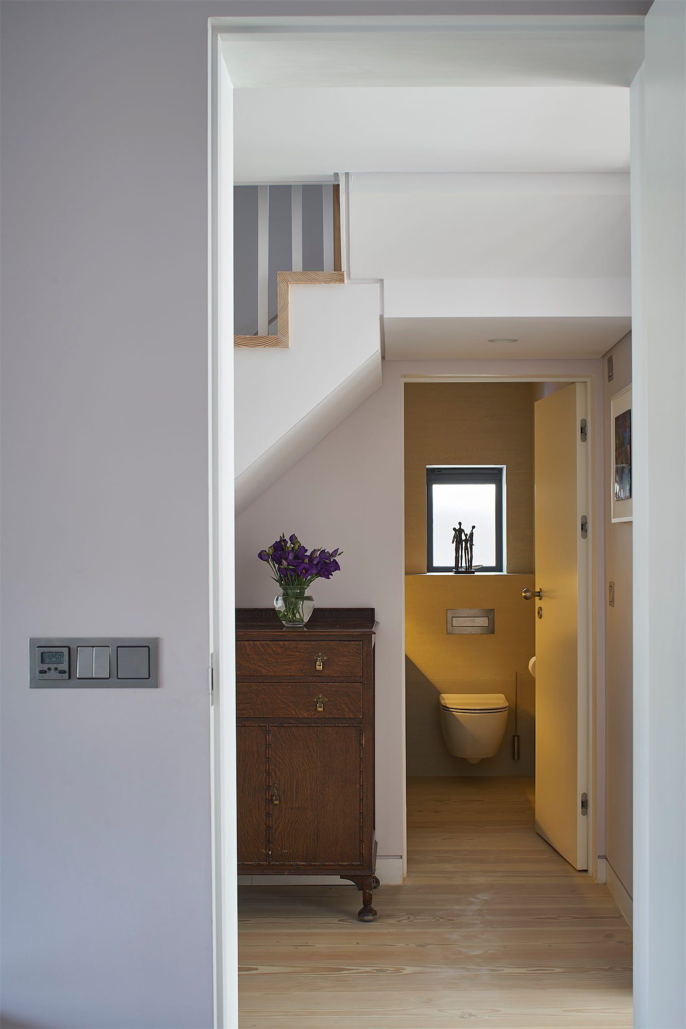 Hallway view into a small bathroom beside a stairway, with a cabinet and purple flowers in the foreground