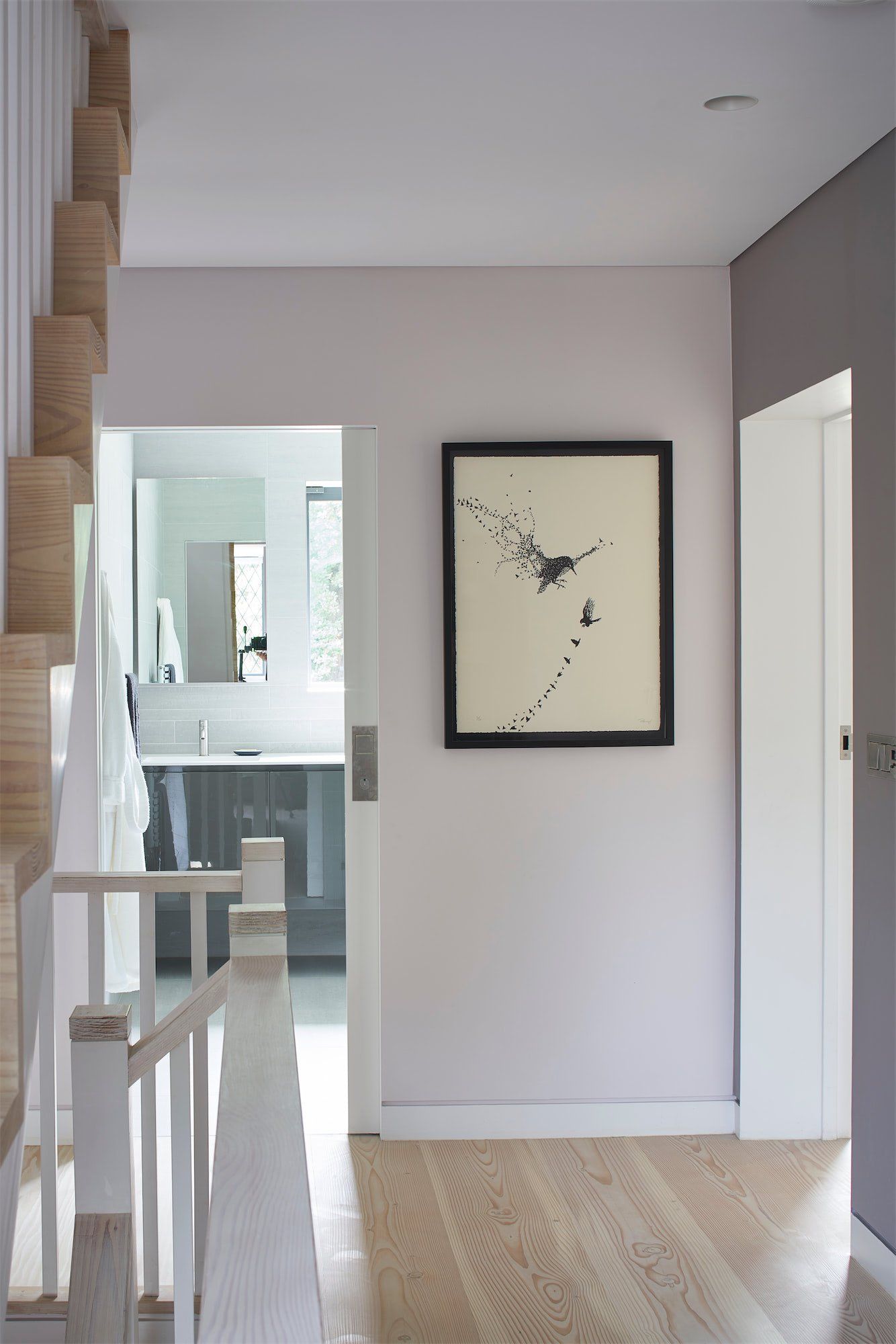 Narrow hallway with pale walls, framed art, and a view into a bright room with a railing and mirror.