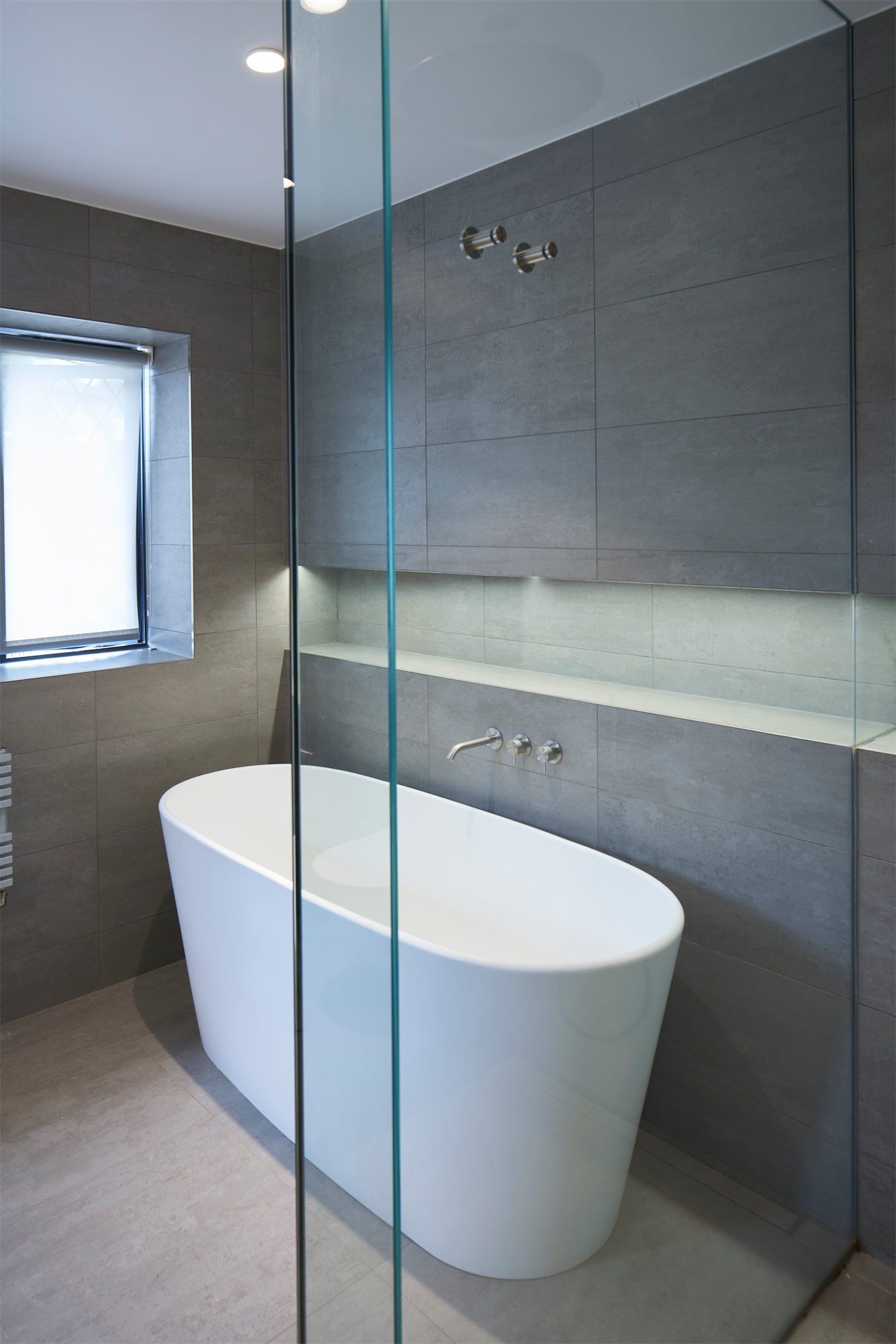 Modern bathroom with a freestanding white tub, glass shower partition, and gray tiled walls.